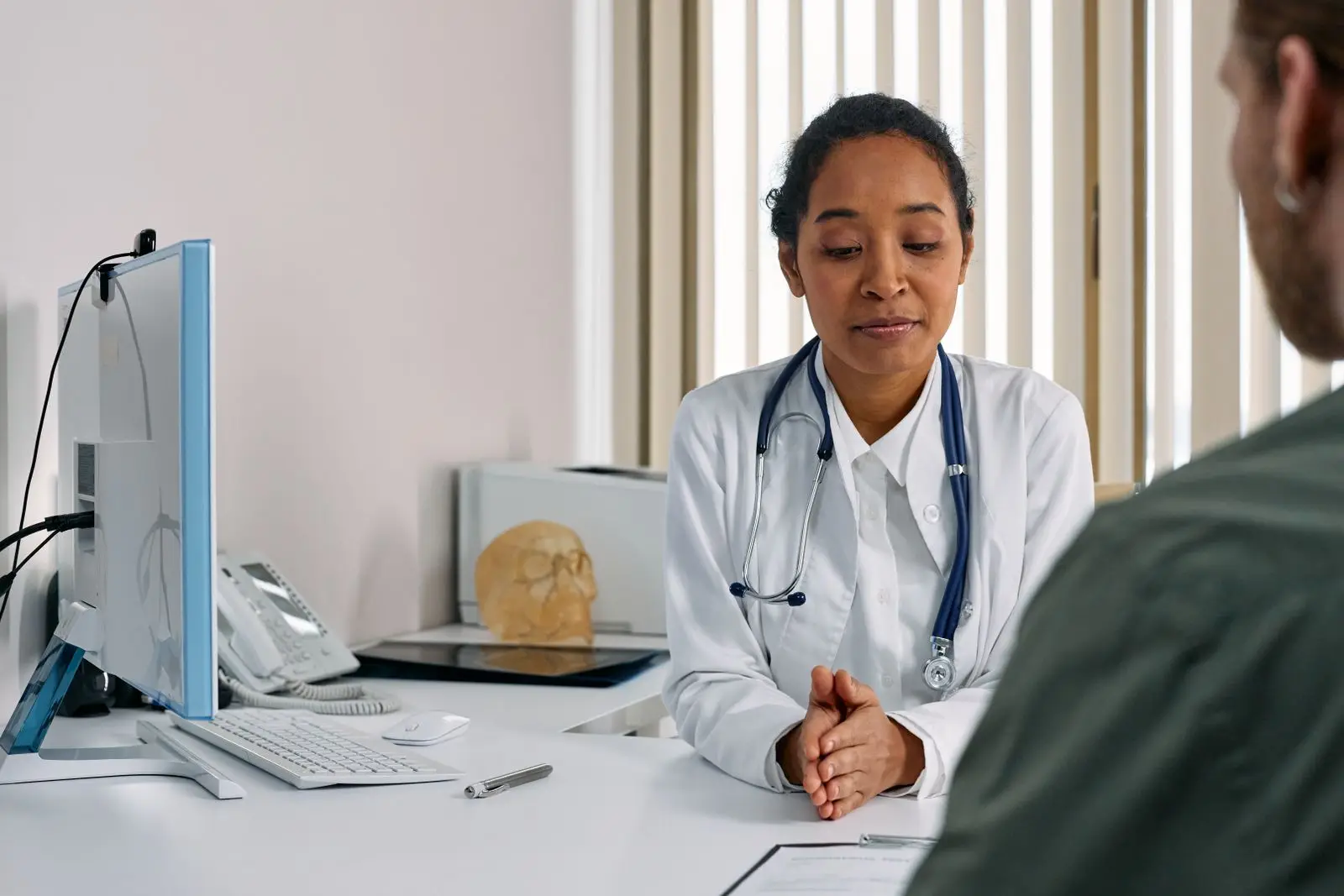 A clinician in a white coat listens to a visitor across her desk.