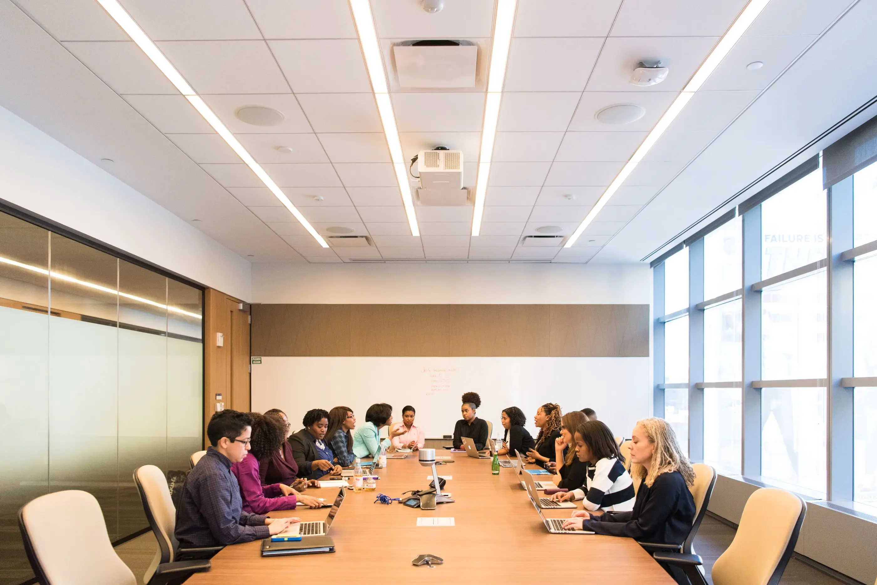 Colleagues work on laptops around a long table in a bright conference room.