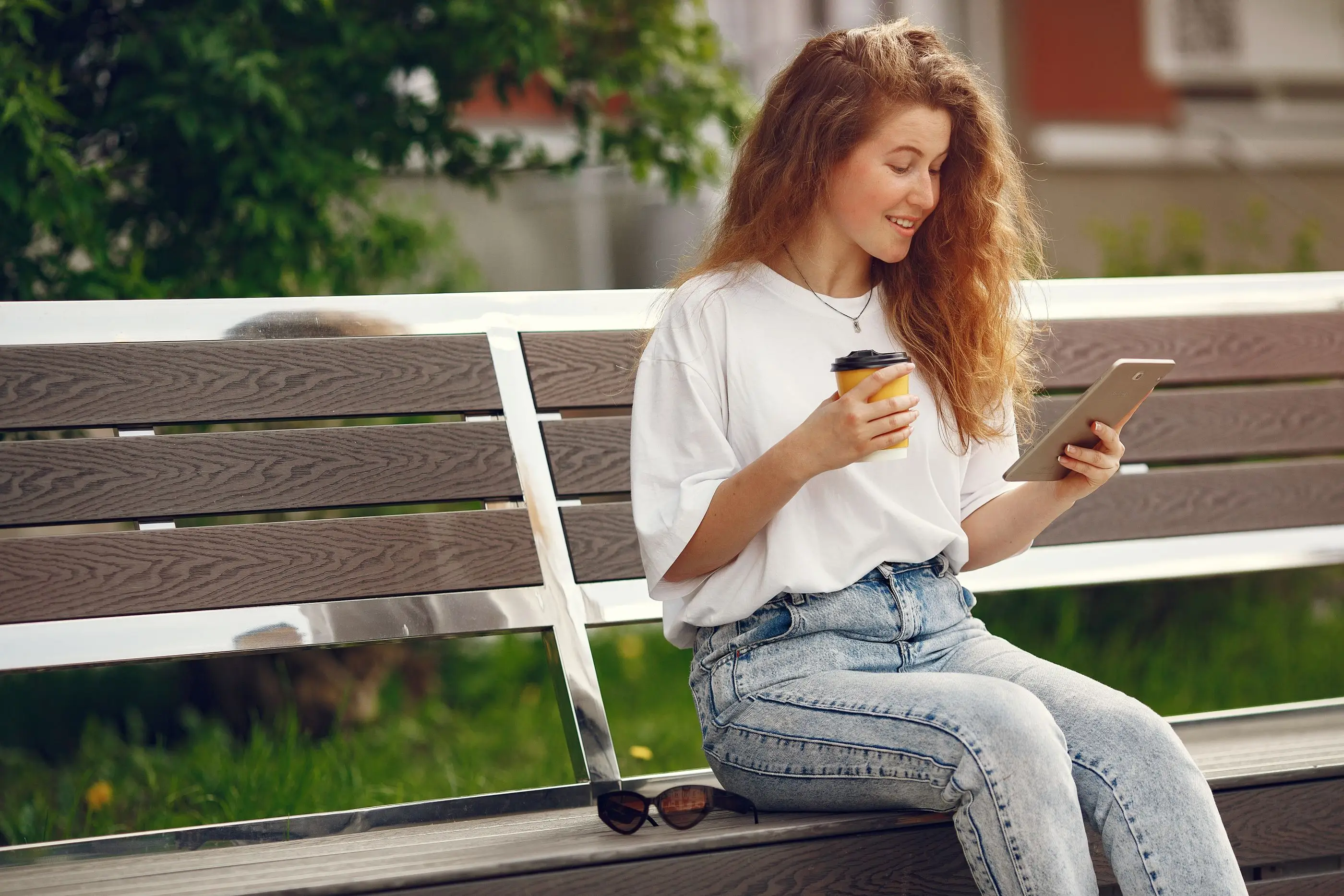 A smiling woman reads on a tablet with a coffee on a sunny park bench.