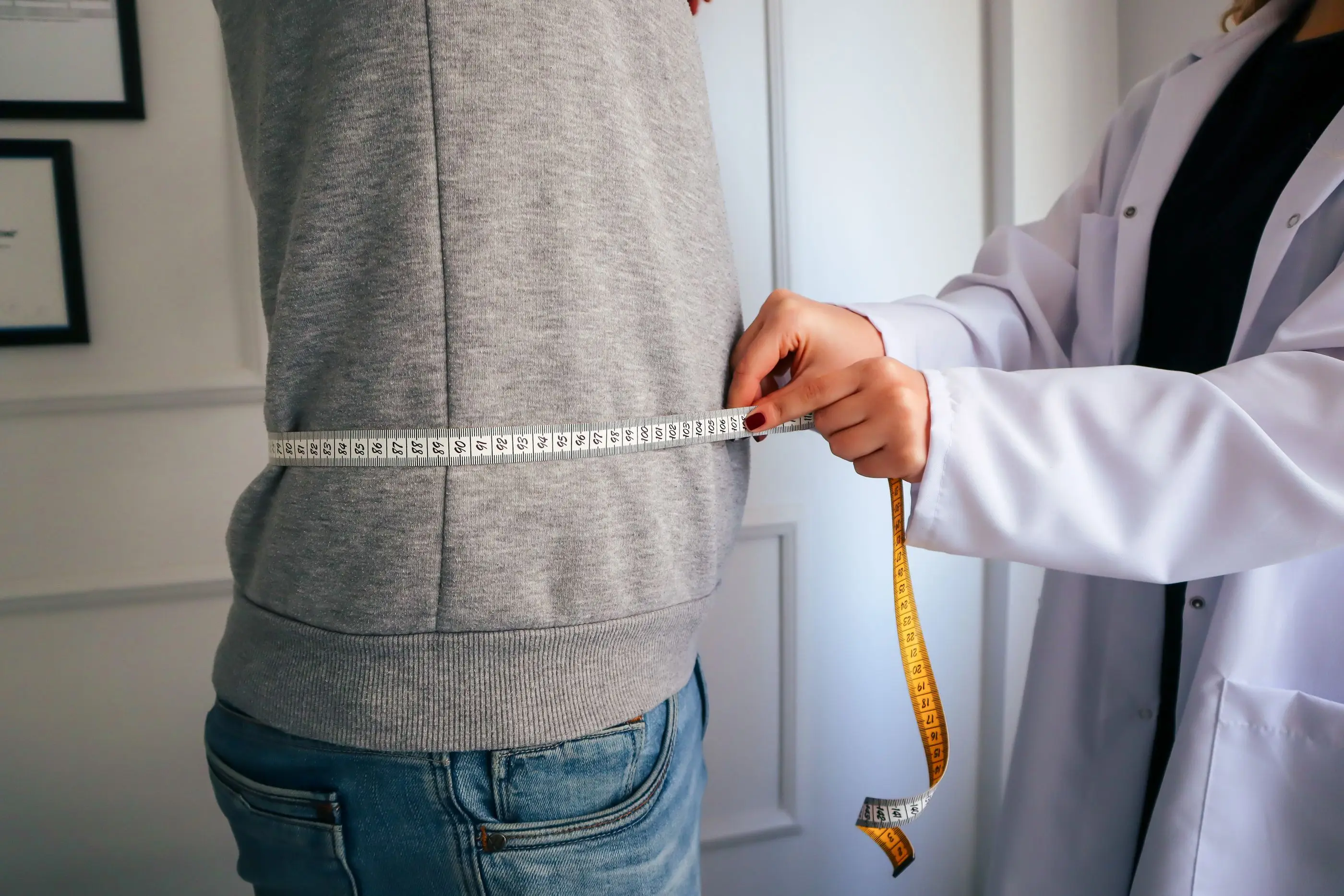 A clinician in a white coat measures a patient's waist with a tape.