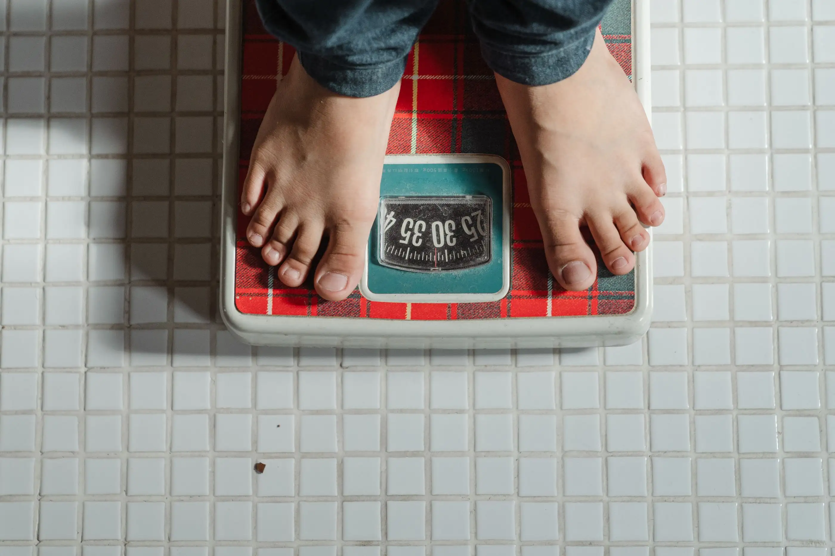 Seen from above, bare feet stand on a bathroom scale on a tiled floor.