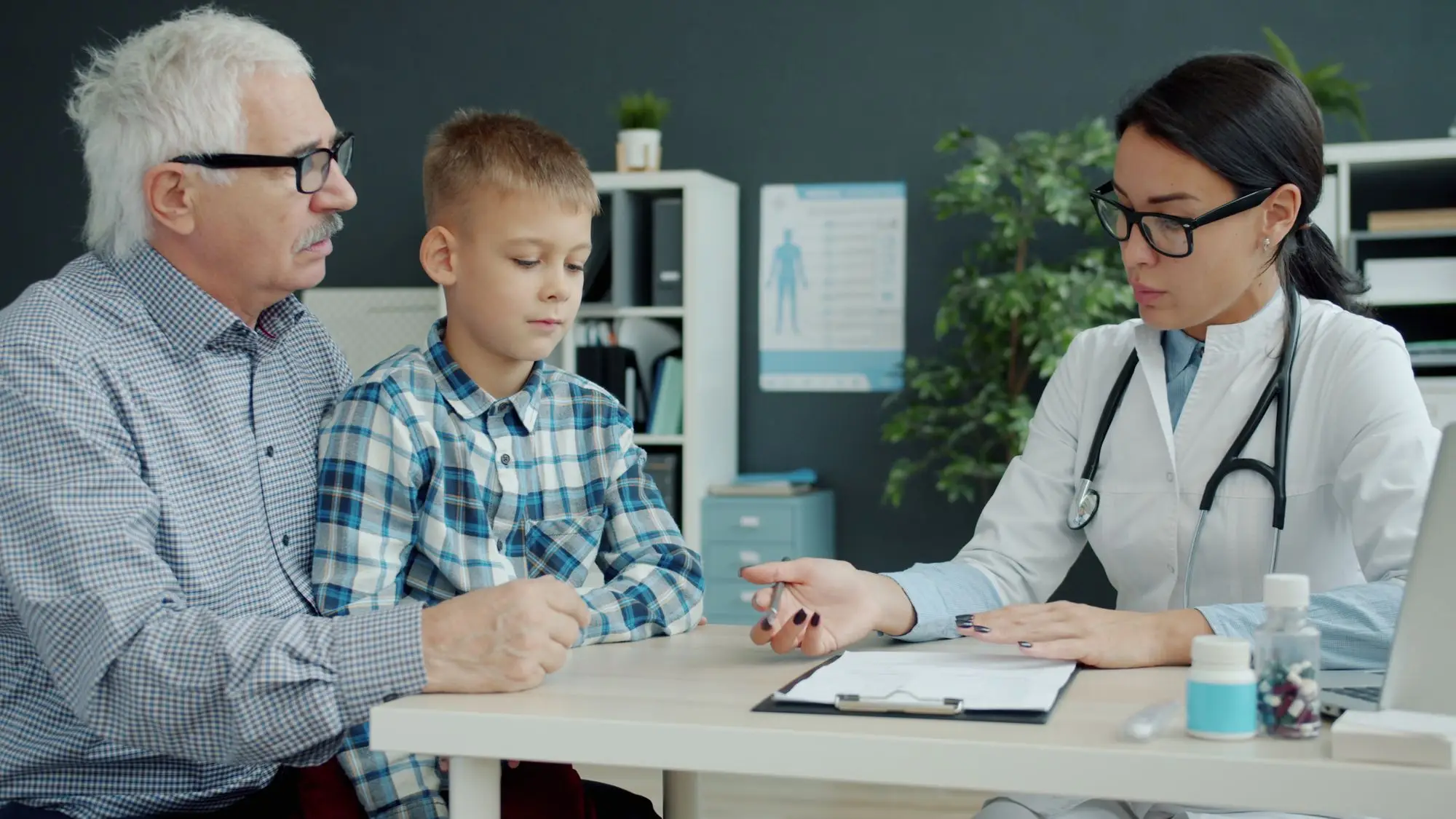 A pharmacist explains a medicine to an older man and a boy at a bright consultation table.