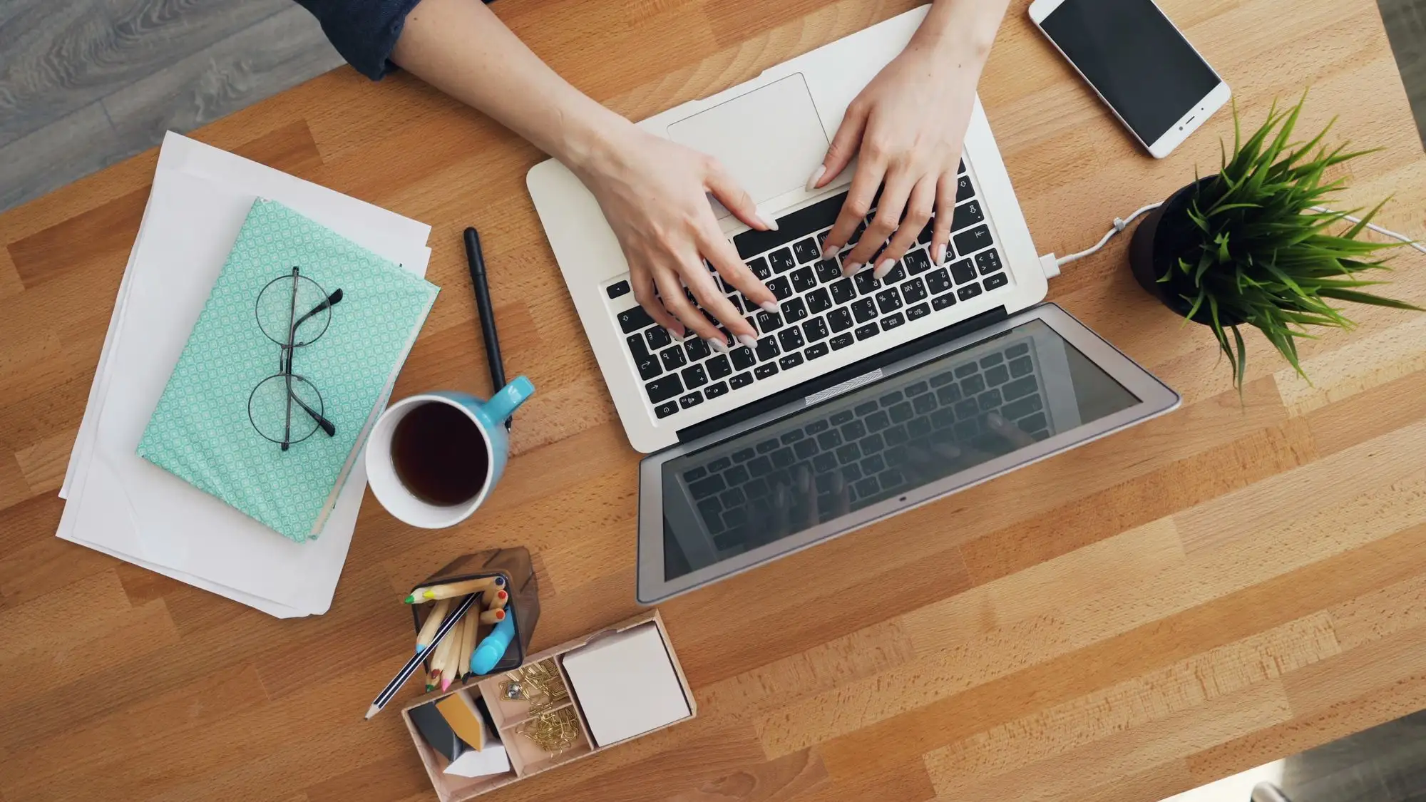Seen from above, hands type on a laptop beside notes, glasses and coffee.