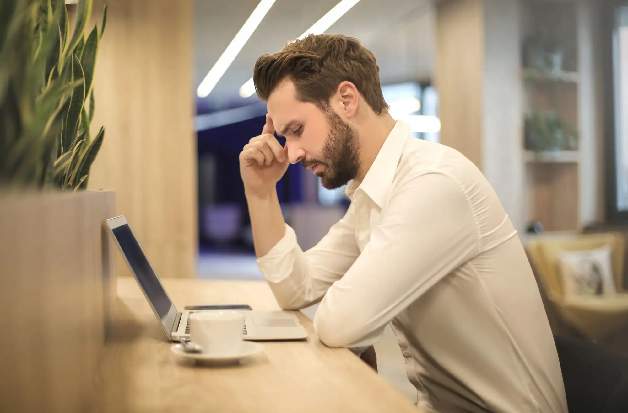 A man rests his head in his hand at a desk beside his laptop.
