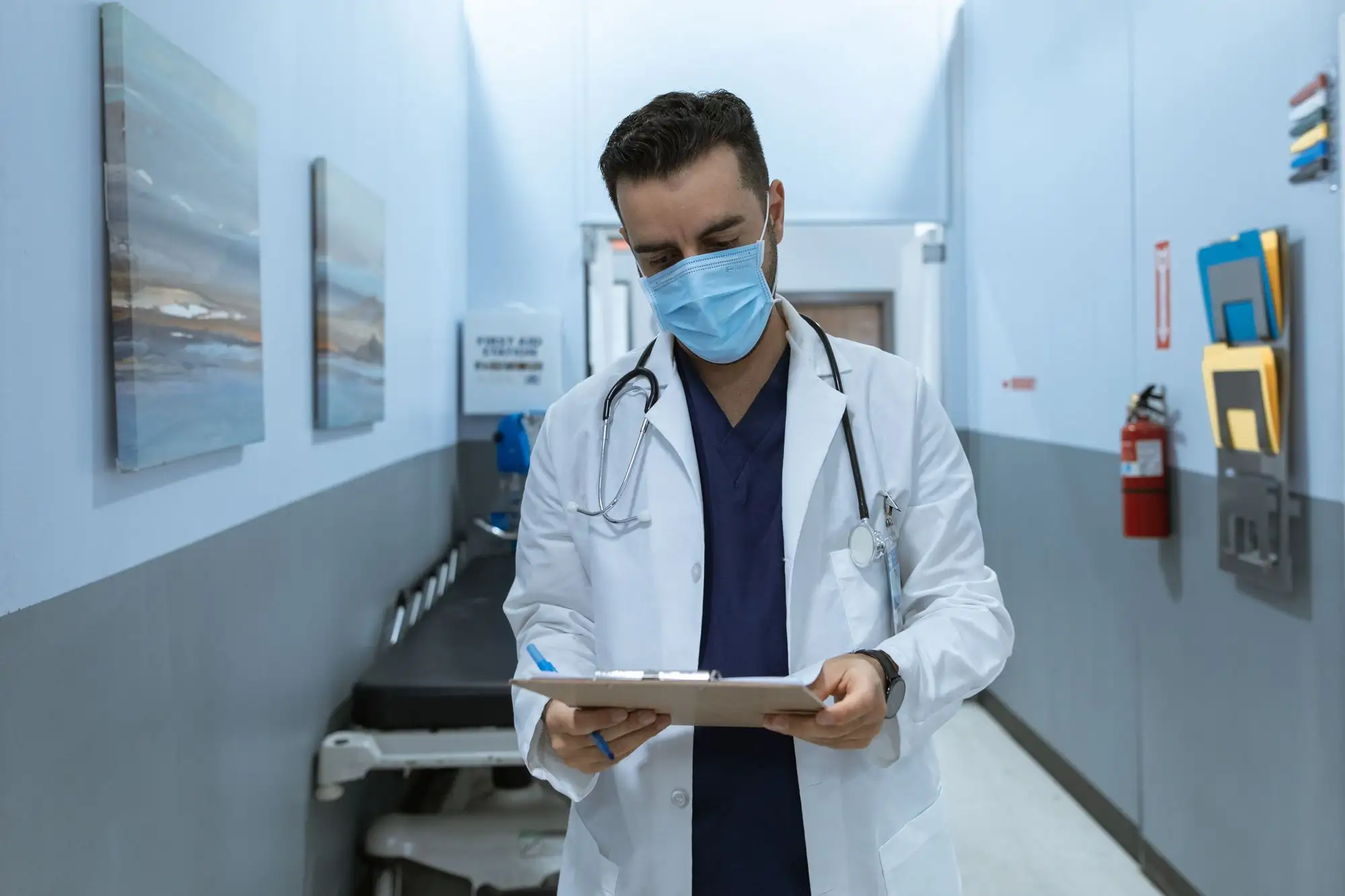 A physician reads a clipboard in a clinic hallway.