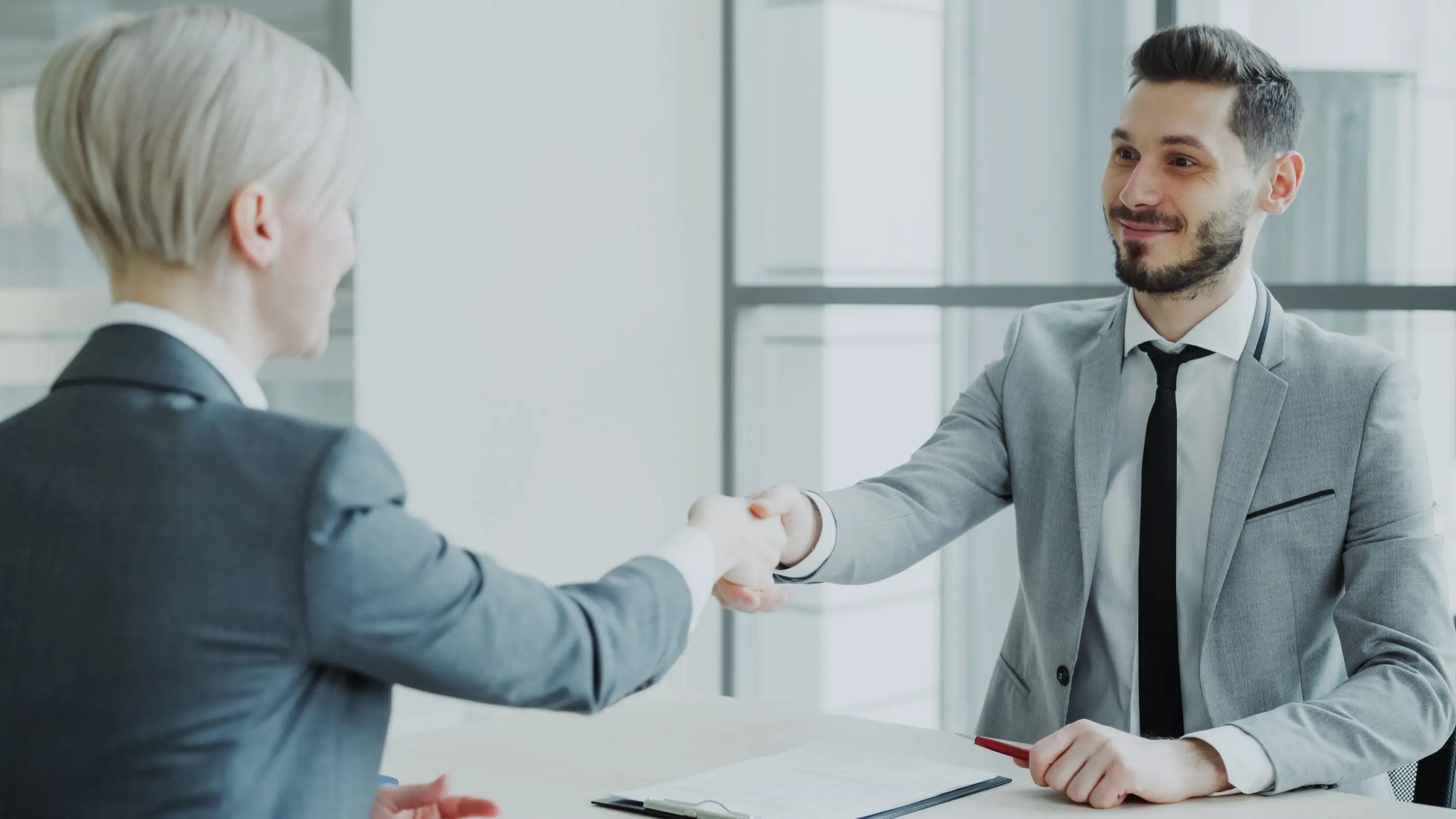 Two people shake hands across a desk at the end of an interview.
