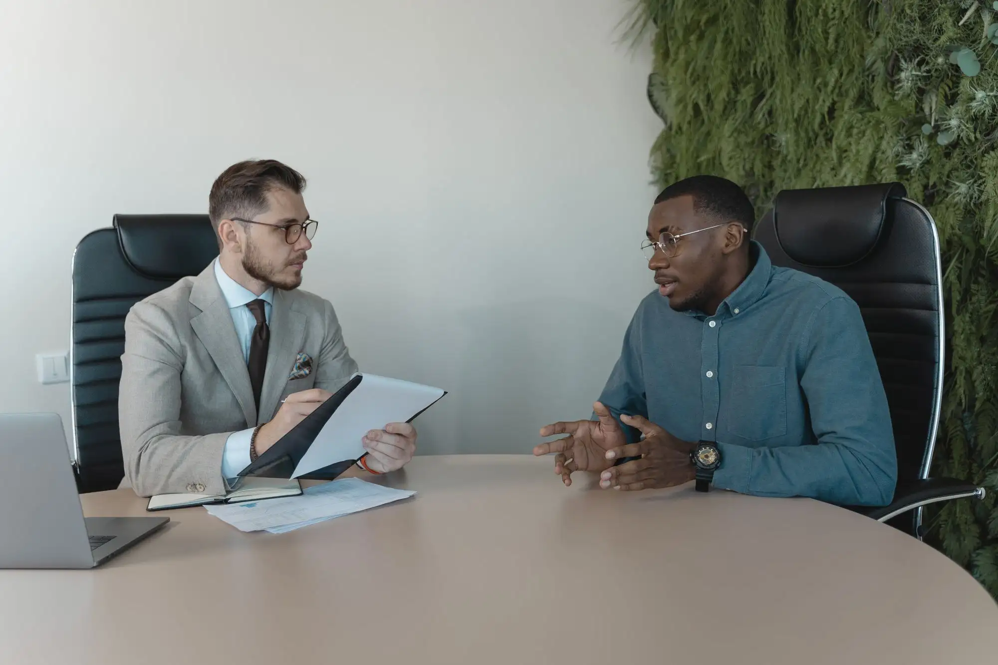 Two men talk across a meeting table, one holding notes.