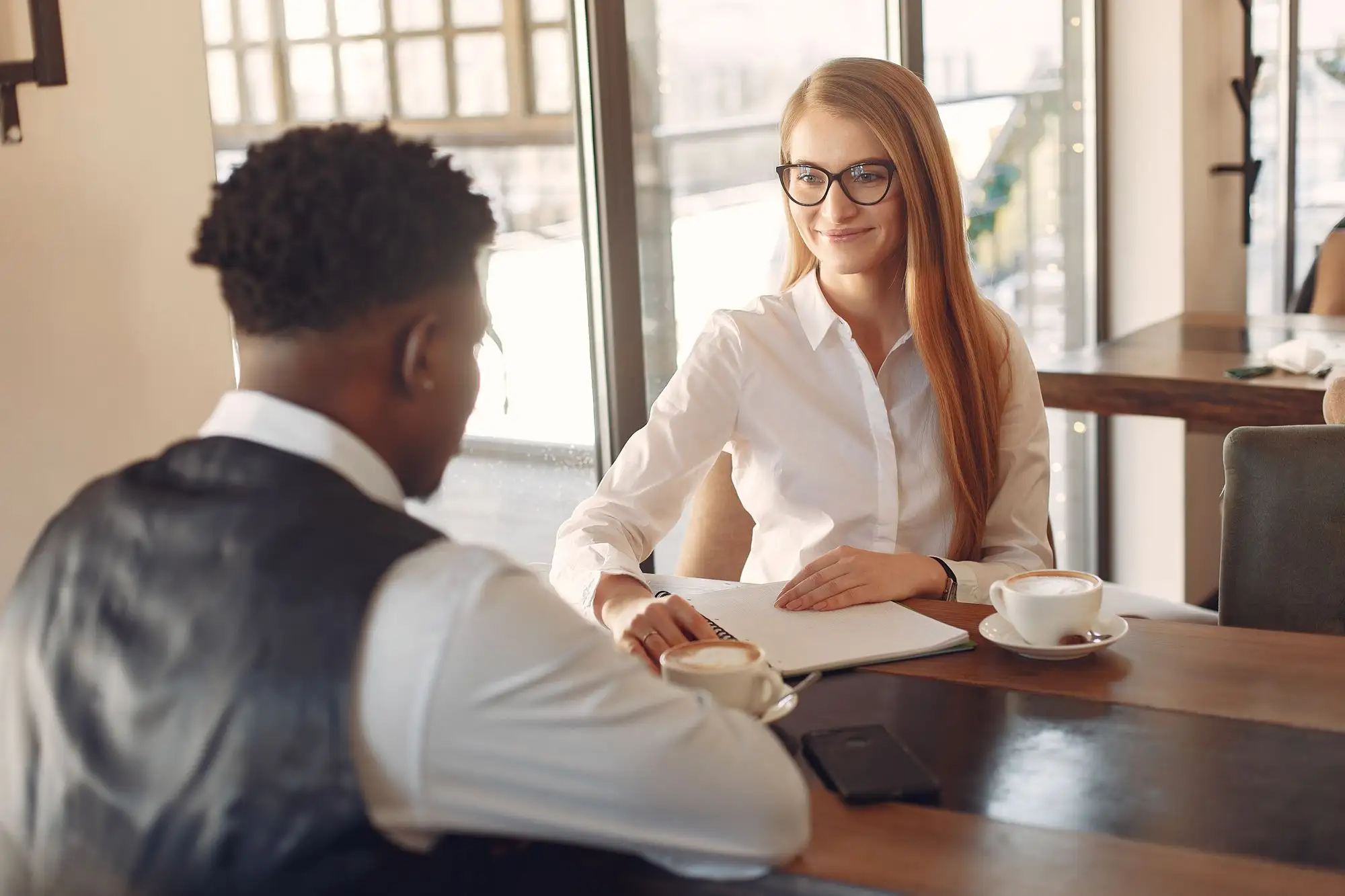 A man and a woman talk over coffee at a table by the window.
