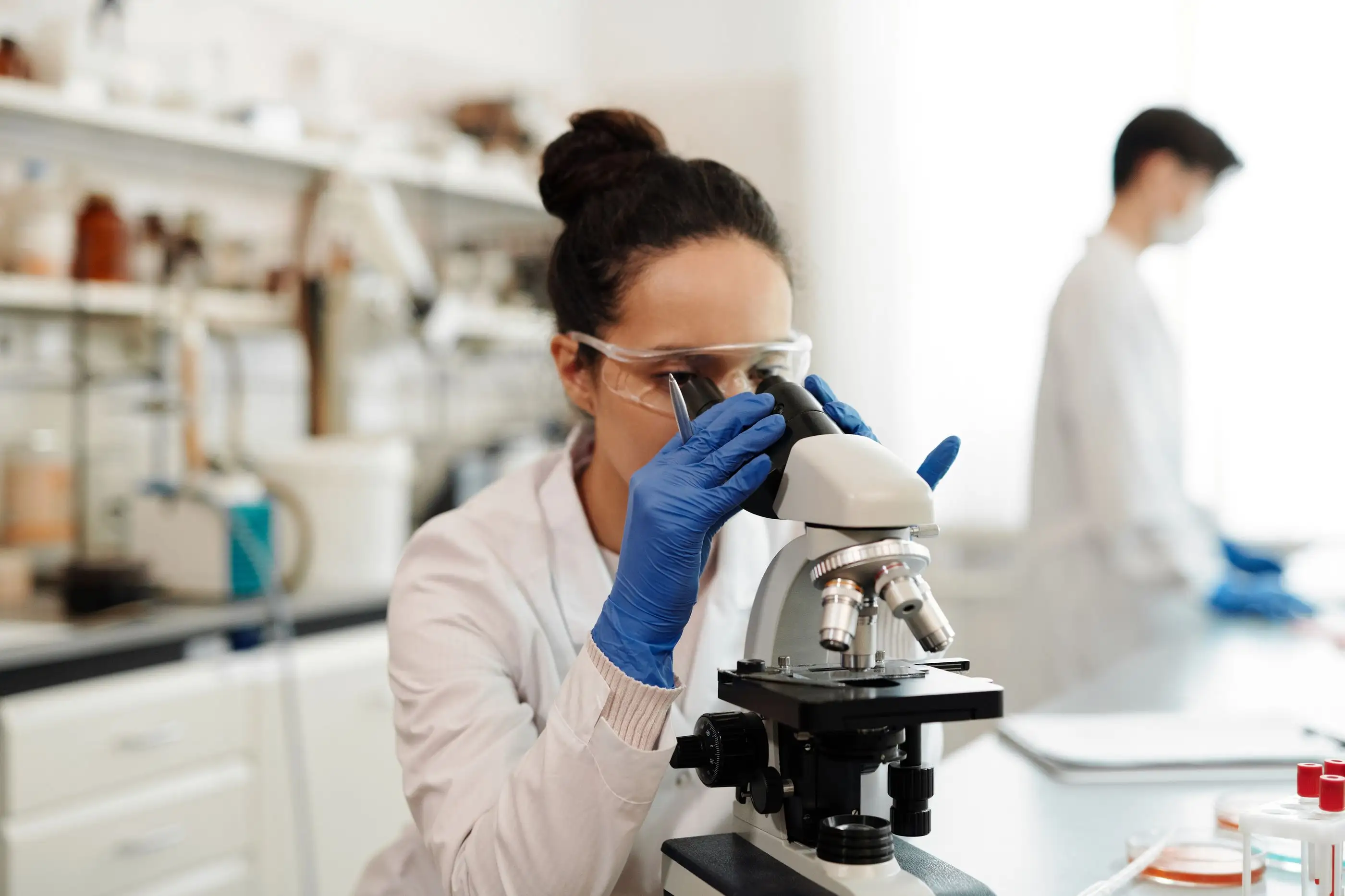 A scientist looks through a microscope while a colleague works behind her.
