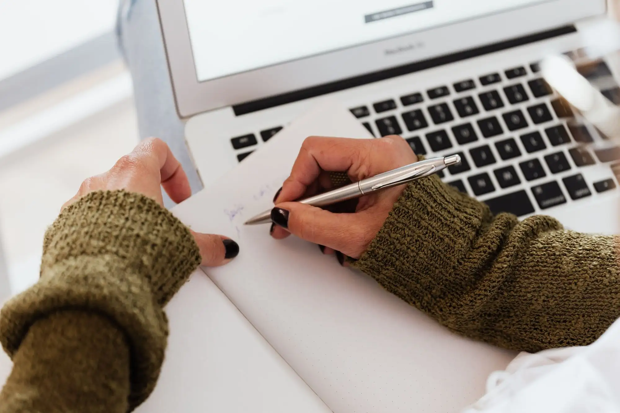 A hand writes notes beside an open laptop.