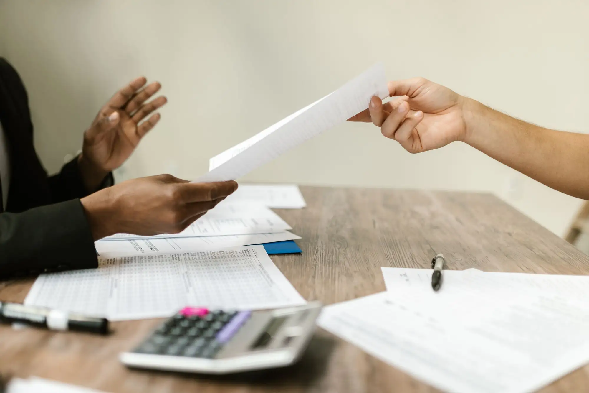 Hands pass a printed document across a desk beside a calculator.
