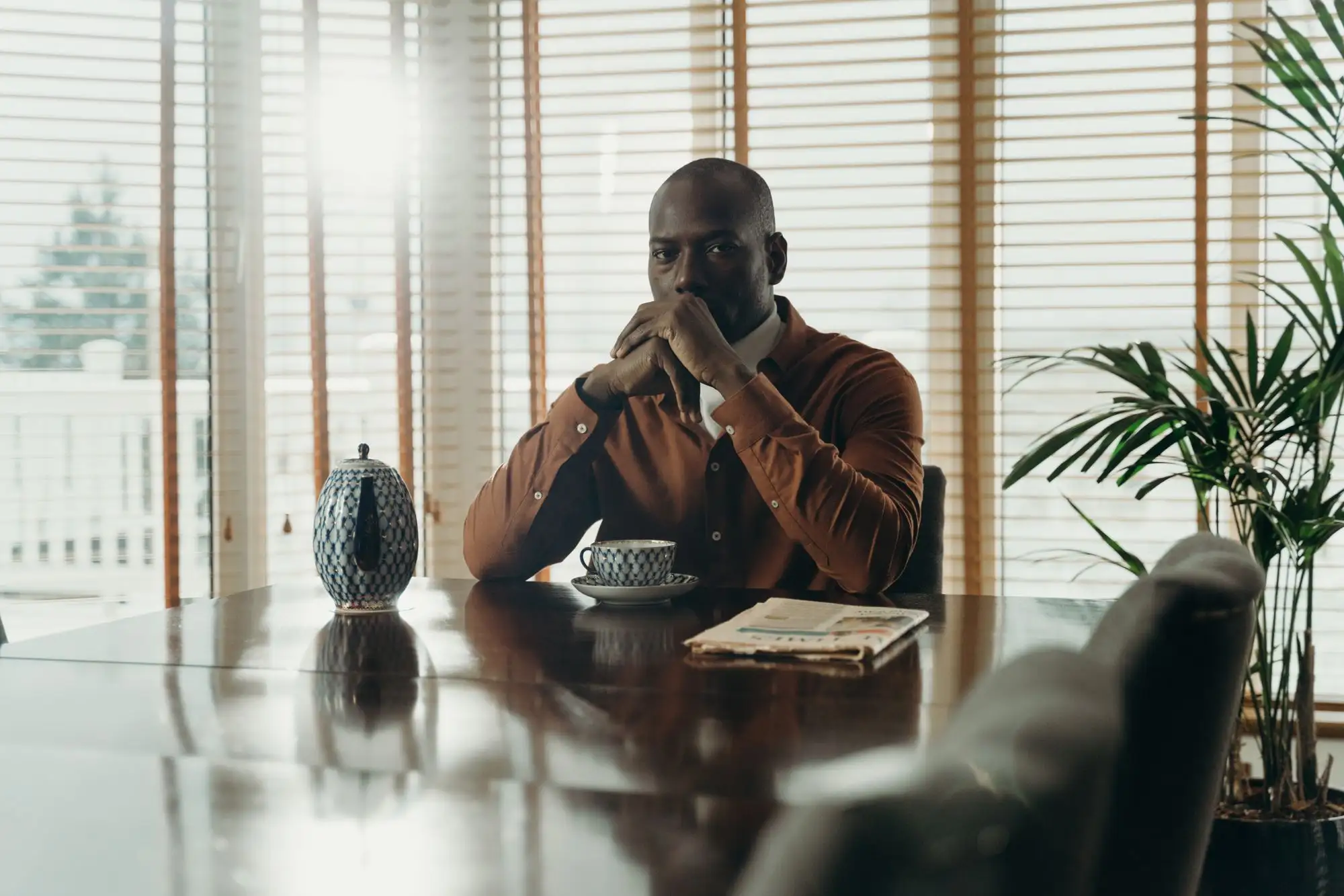 A man sits at a long table, thinking, with a cup of coffee.