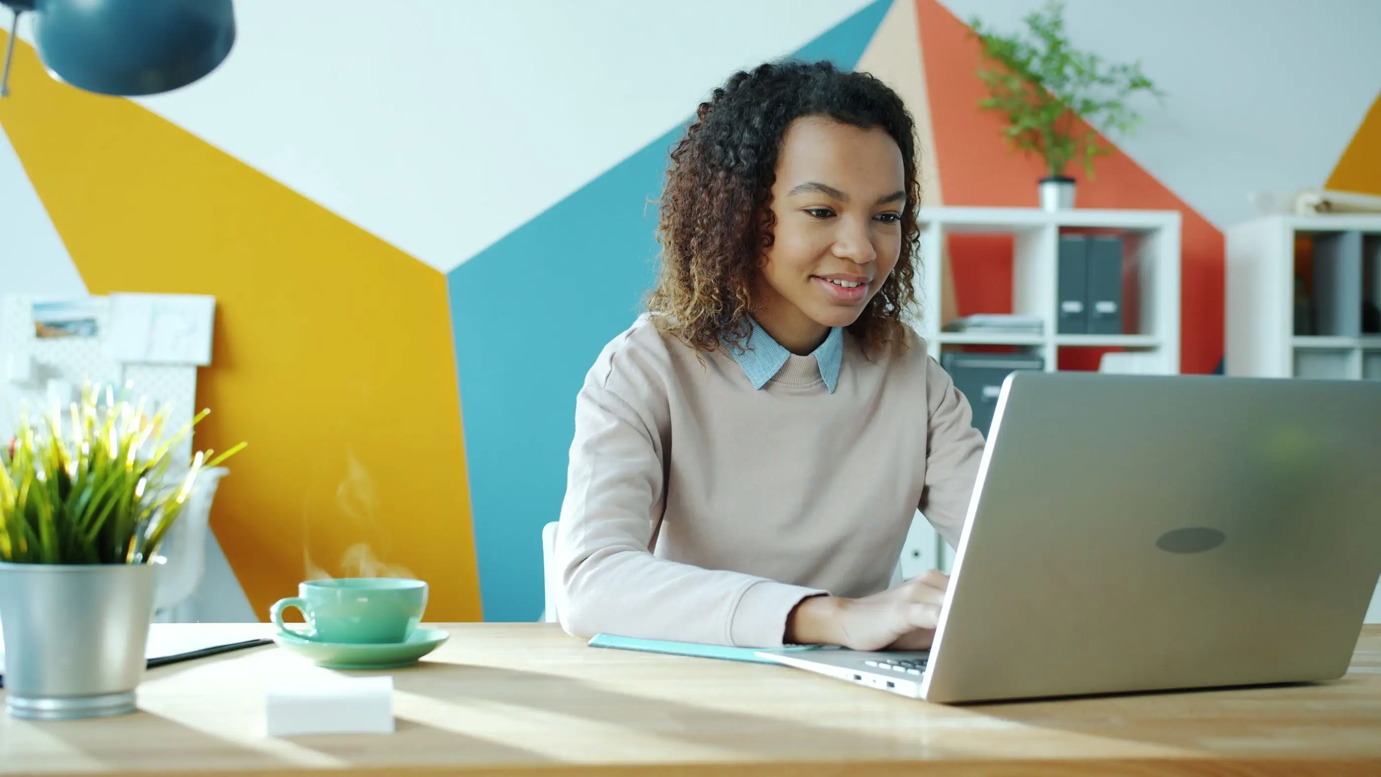 A smiling young woman works on her laptop in a bright, colorful office.