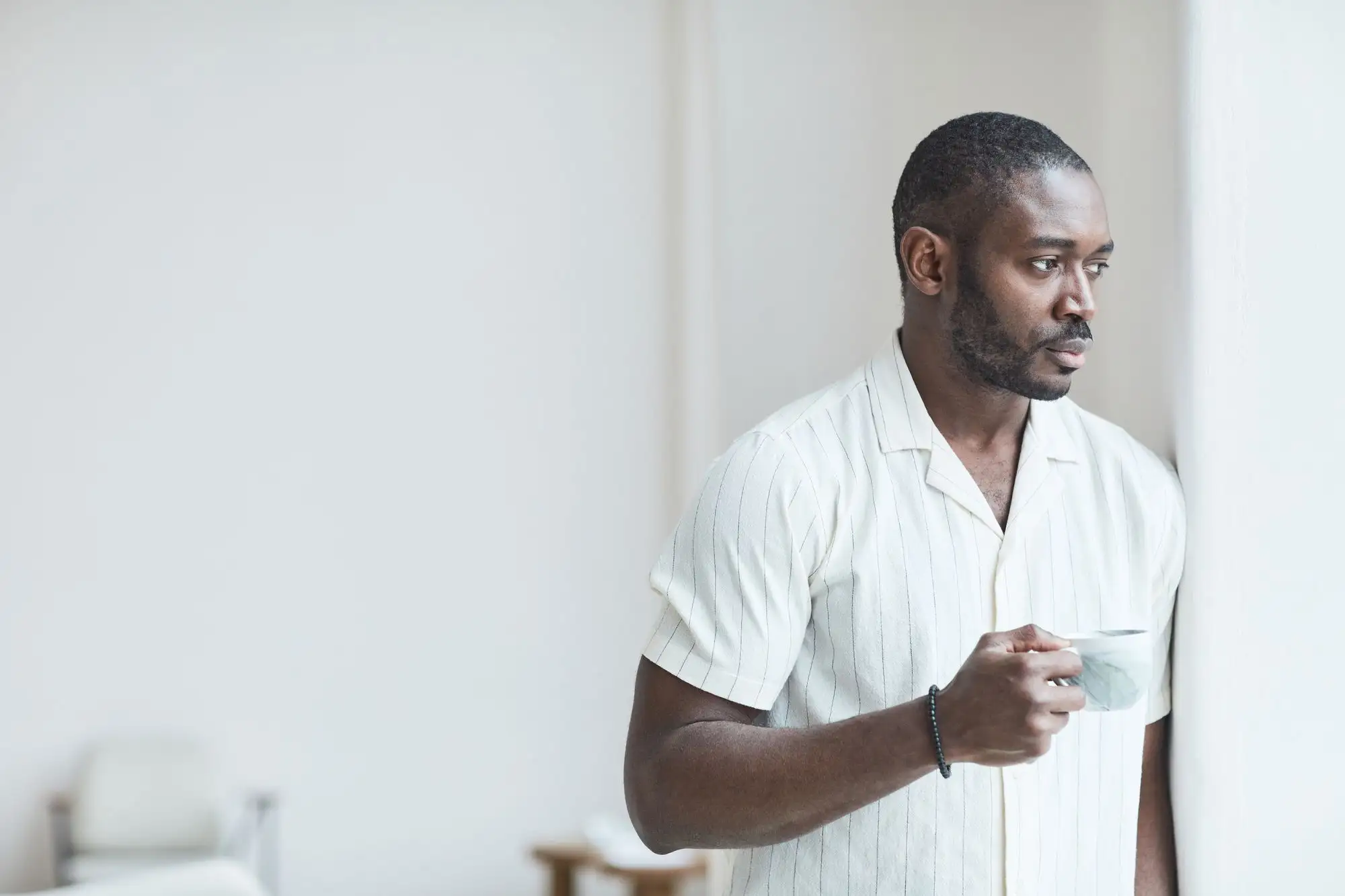 A man holds a cup and looks thoughtfully out of a window.