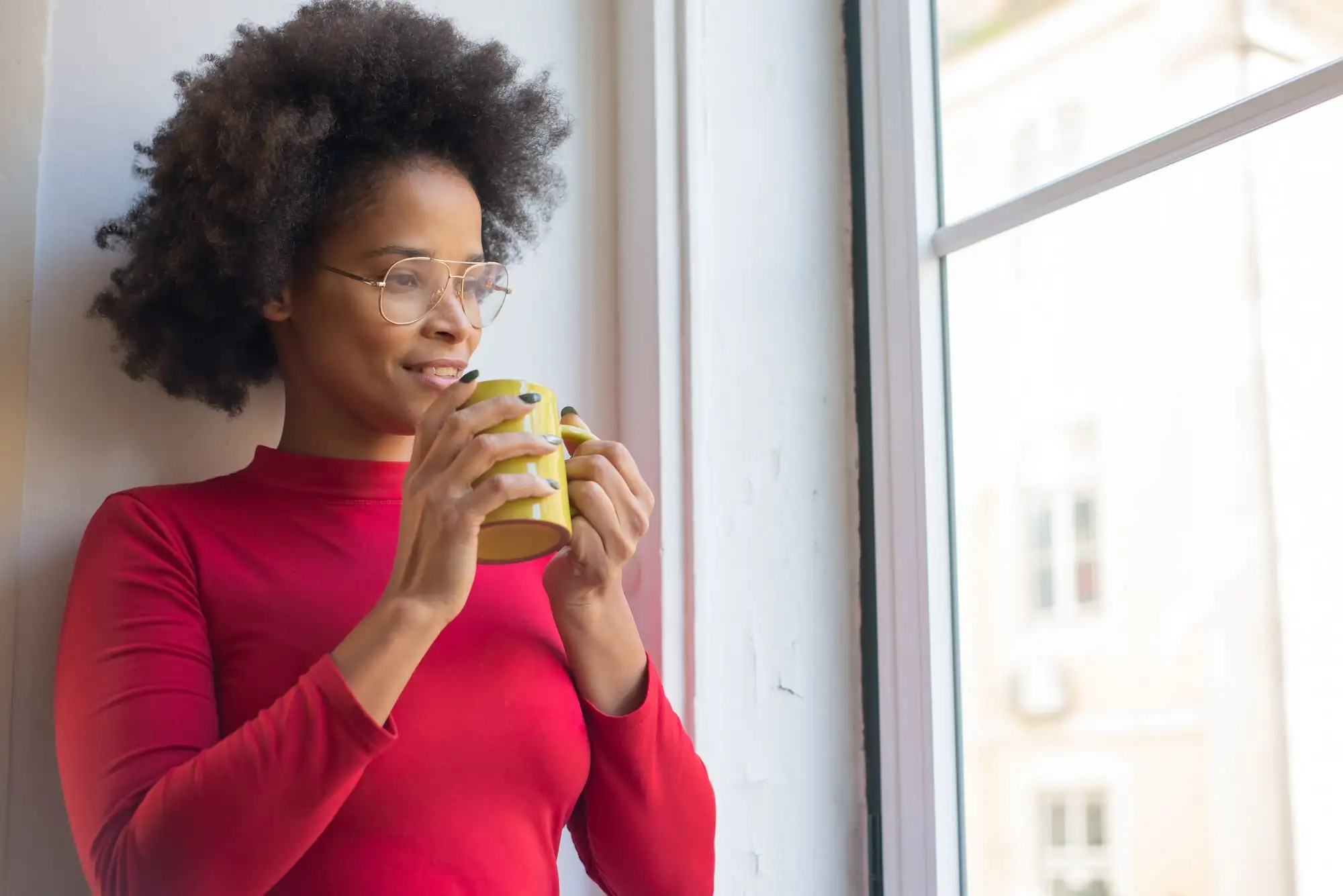A woman in a red top holds a mug and looks out of a window, thinking.