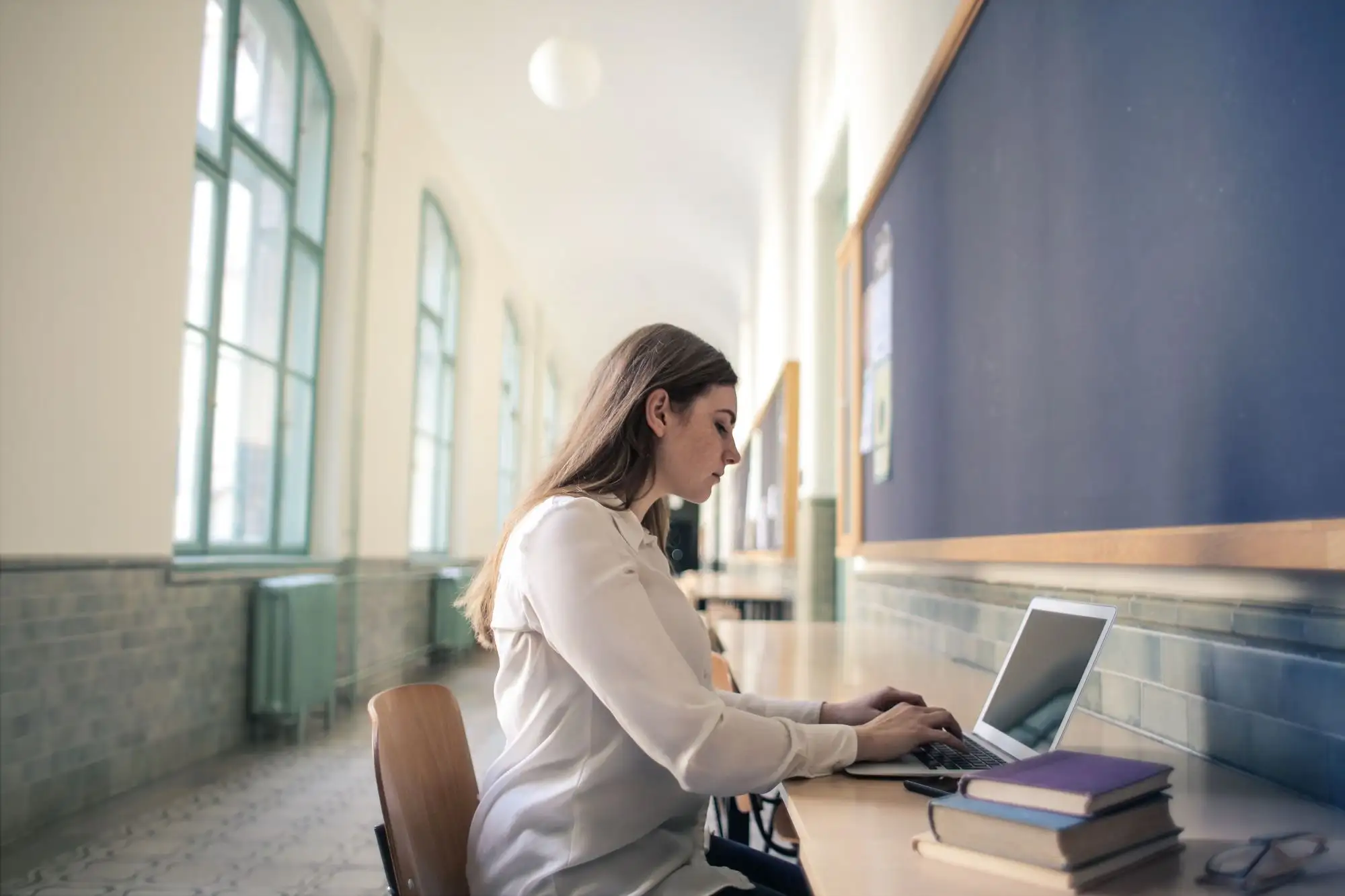A woman works on a laptop at a long desk in a sunny hallway.