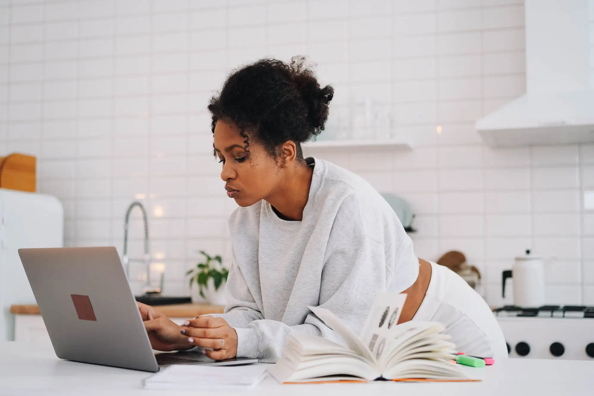 A woman reads and takes notes at her laptop in a bright kitchen.