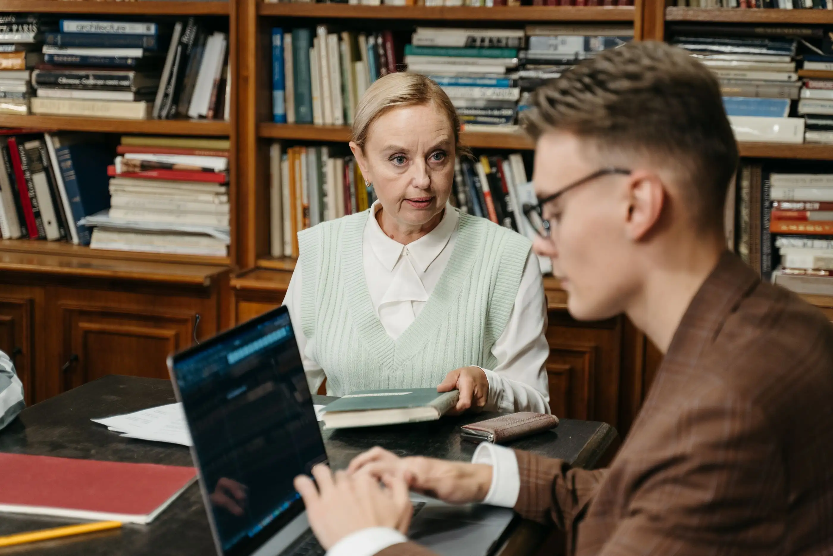 An experienced mentor talks with a young colleague at a desk in a book-lined office.