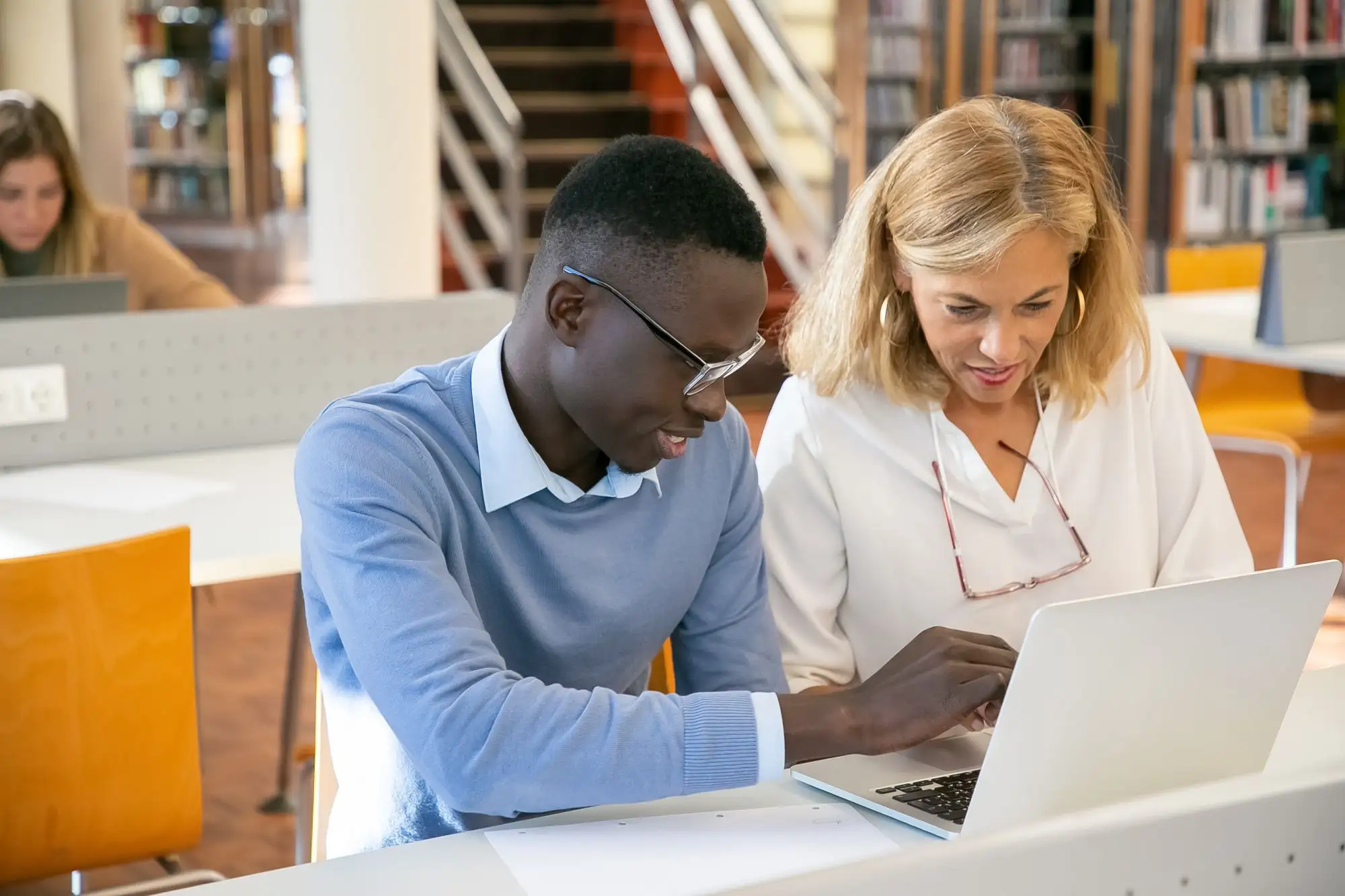 A mentor and a young man review work on a laptop in a library.