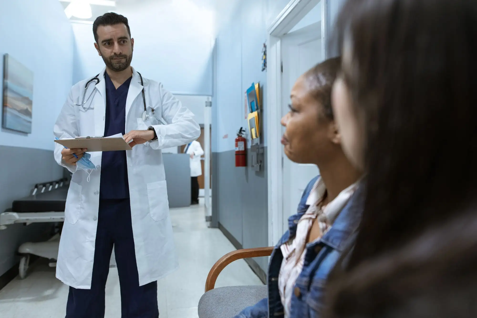 A physician holding a clipboard talks with a student in a clinic hallway.