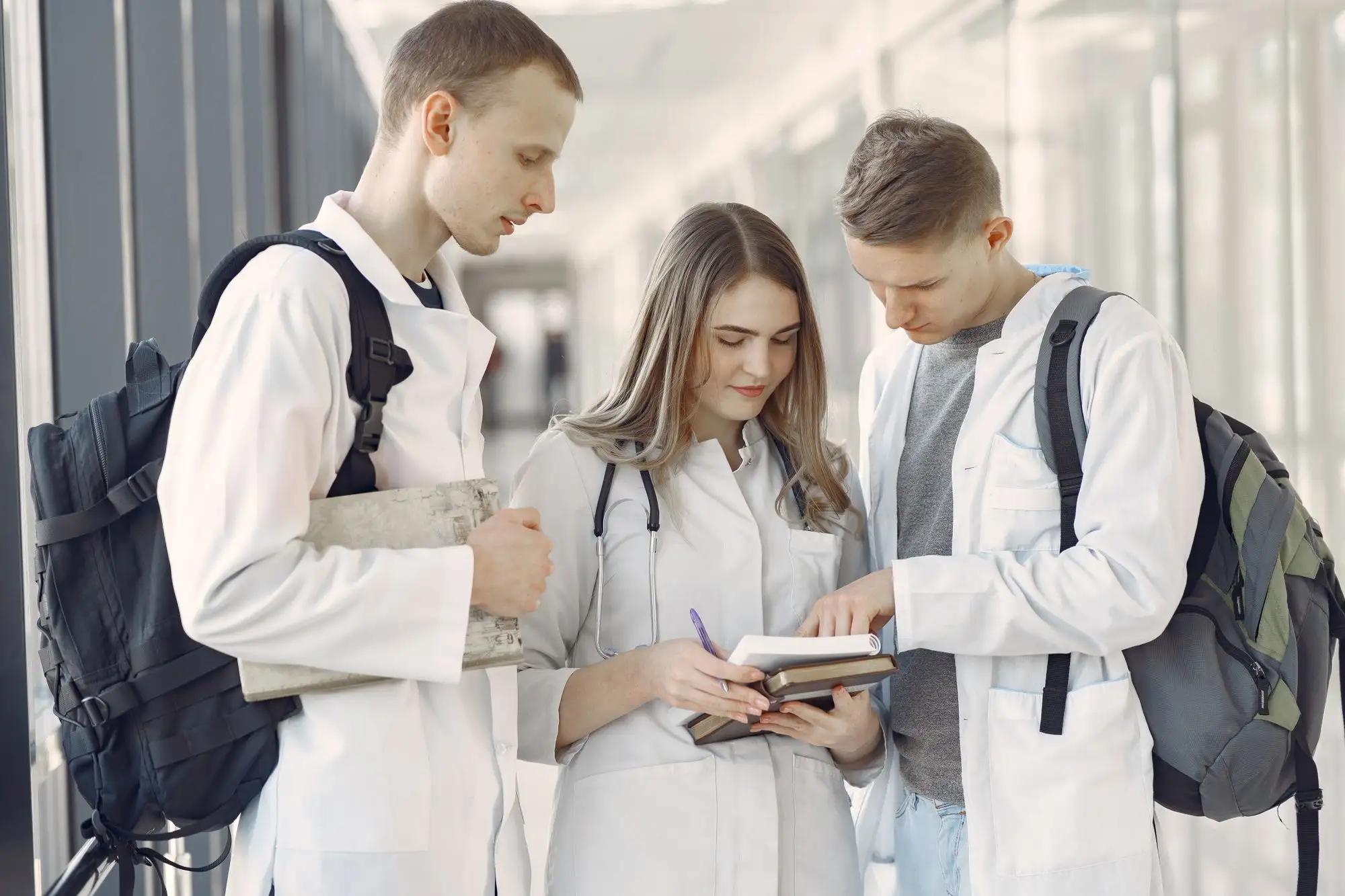 Three students in white coats with backpacks read a notebook together.