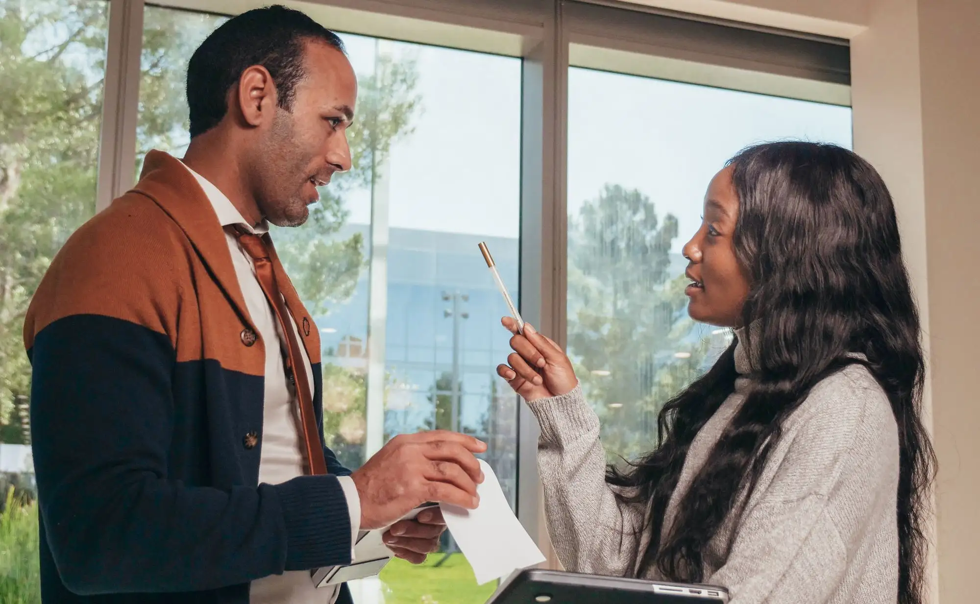 A man and a woman talk by a sunny window, one holding notes.