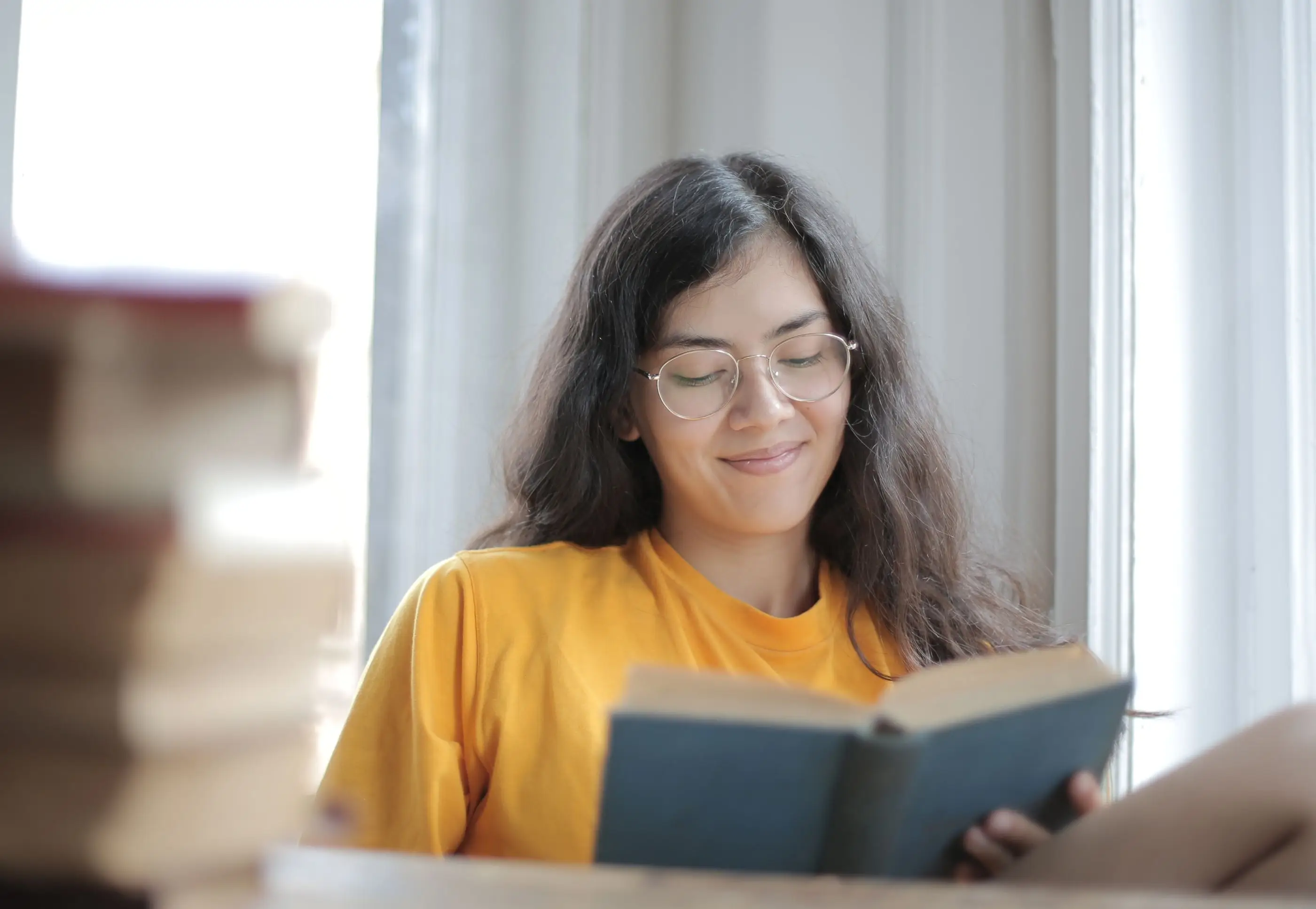 A smiling young woman in glasses reads a book by a bright window.