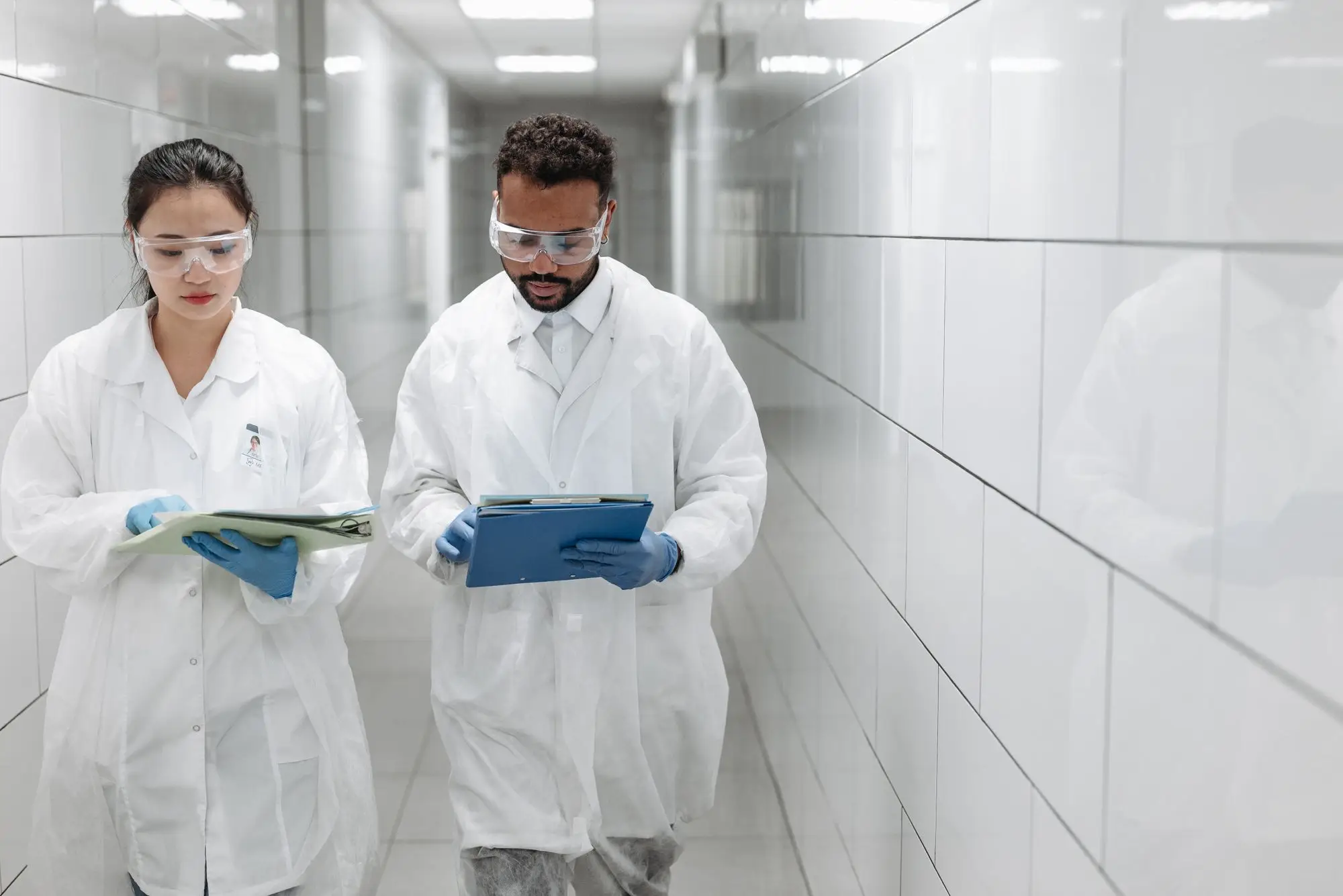 Two scientists in lab coats and safety glasses review clipboards in a bright corridor.