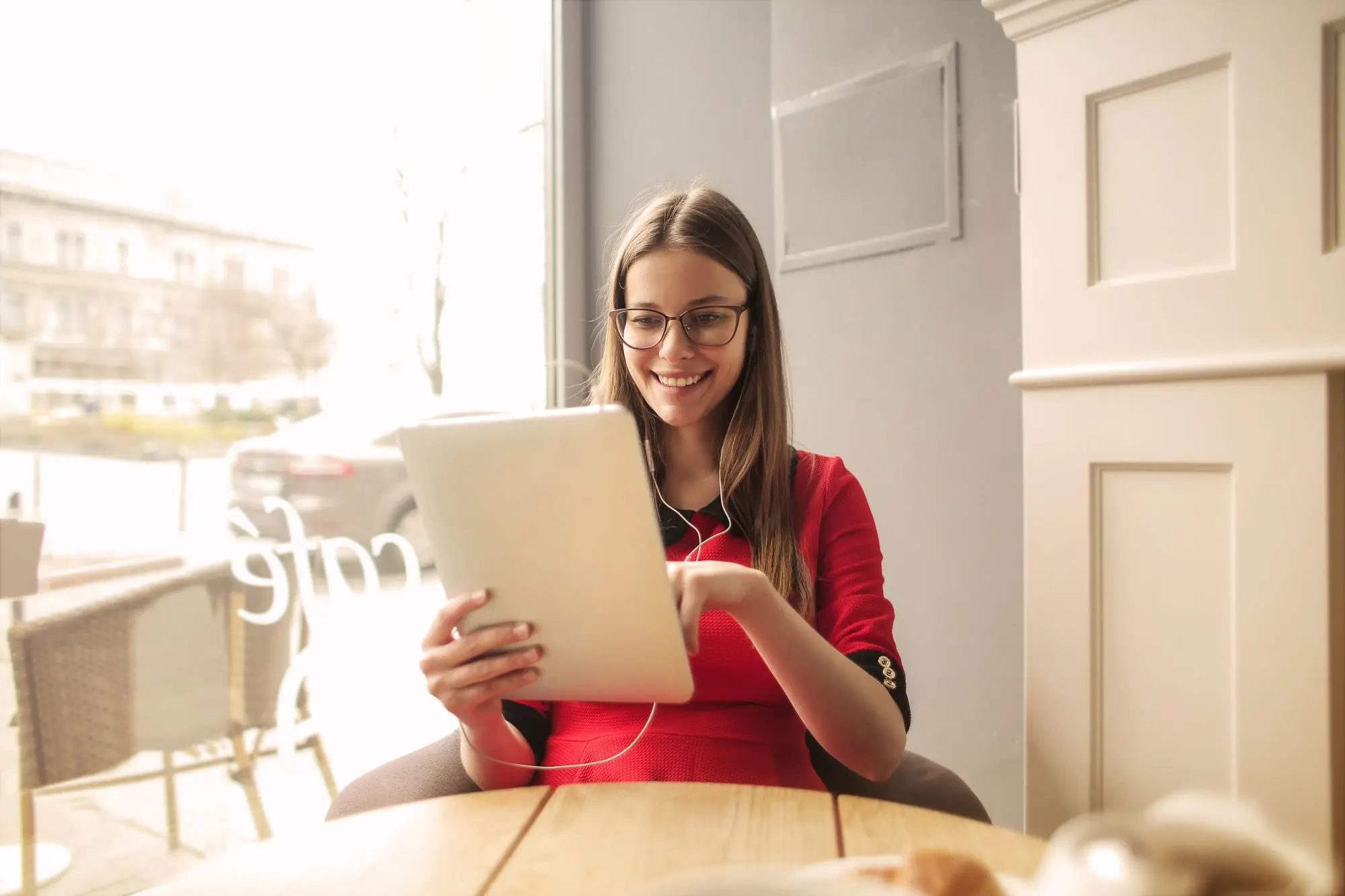 A smiling woman reads on a tablet at a café table by the window.