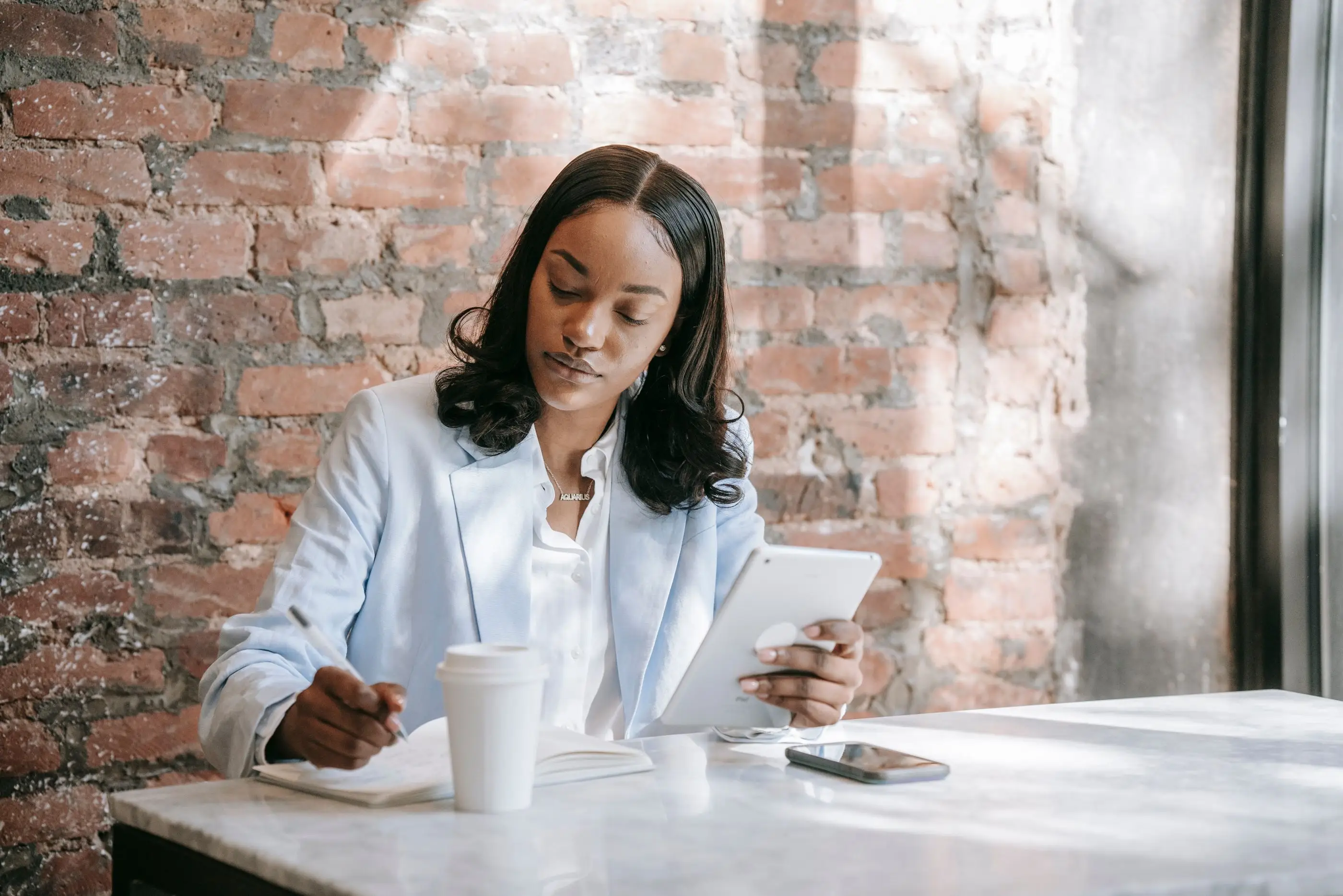 A woman reads on a tablet and takes notes at a sunlit table.