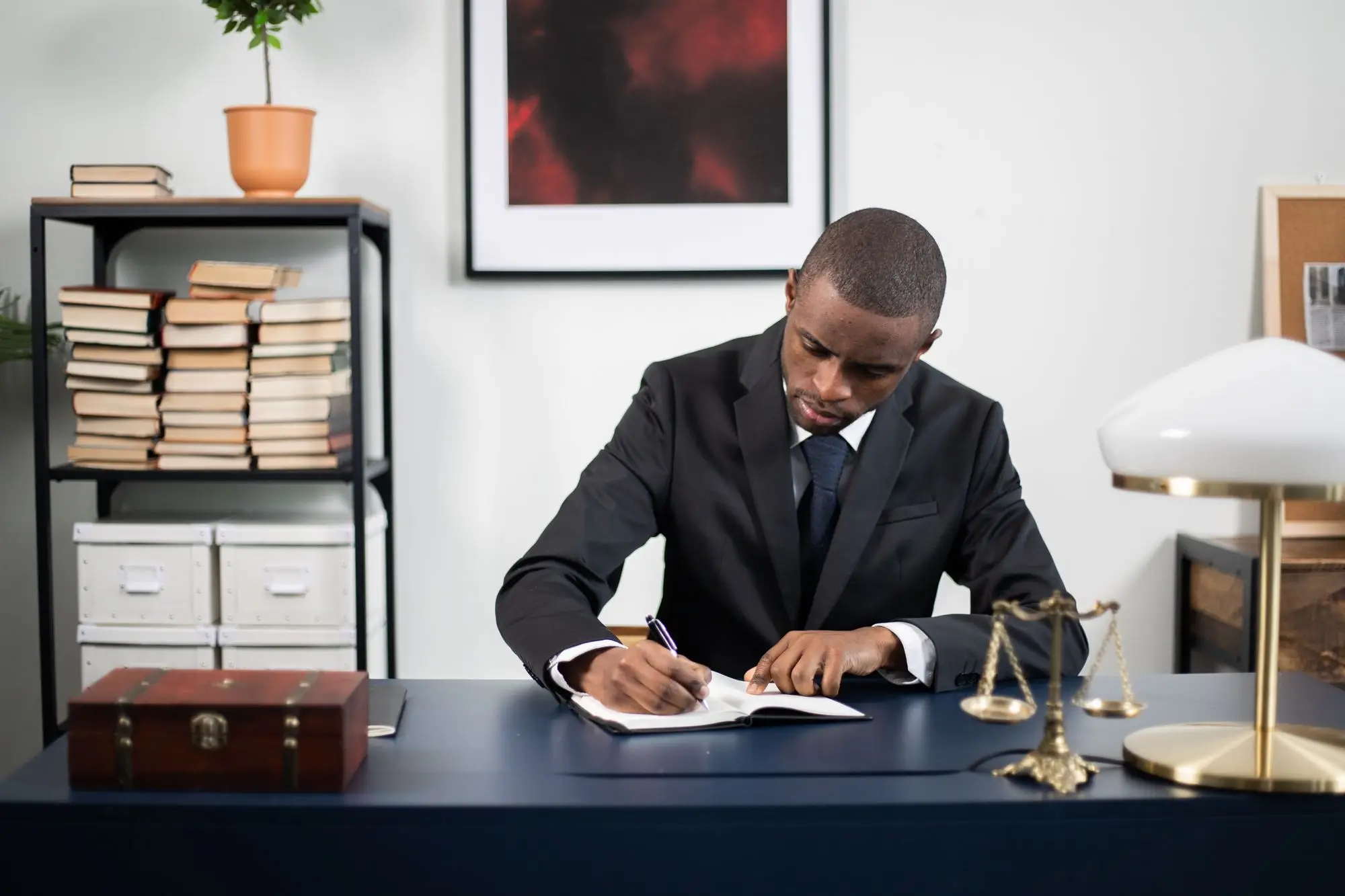 A man in a suit writes at a desk beside a set of brass scales.