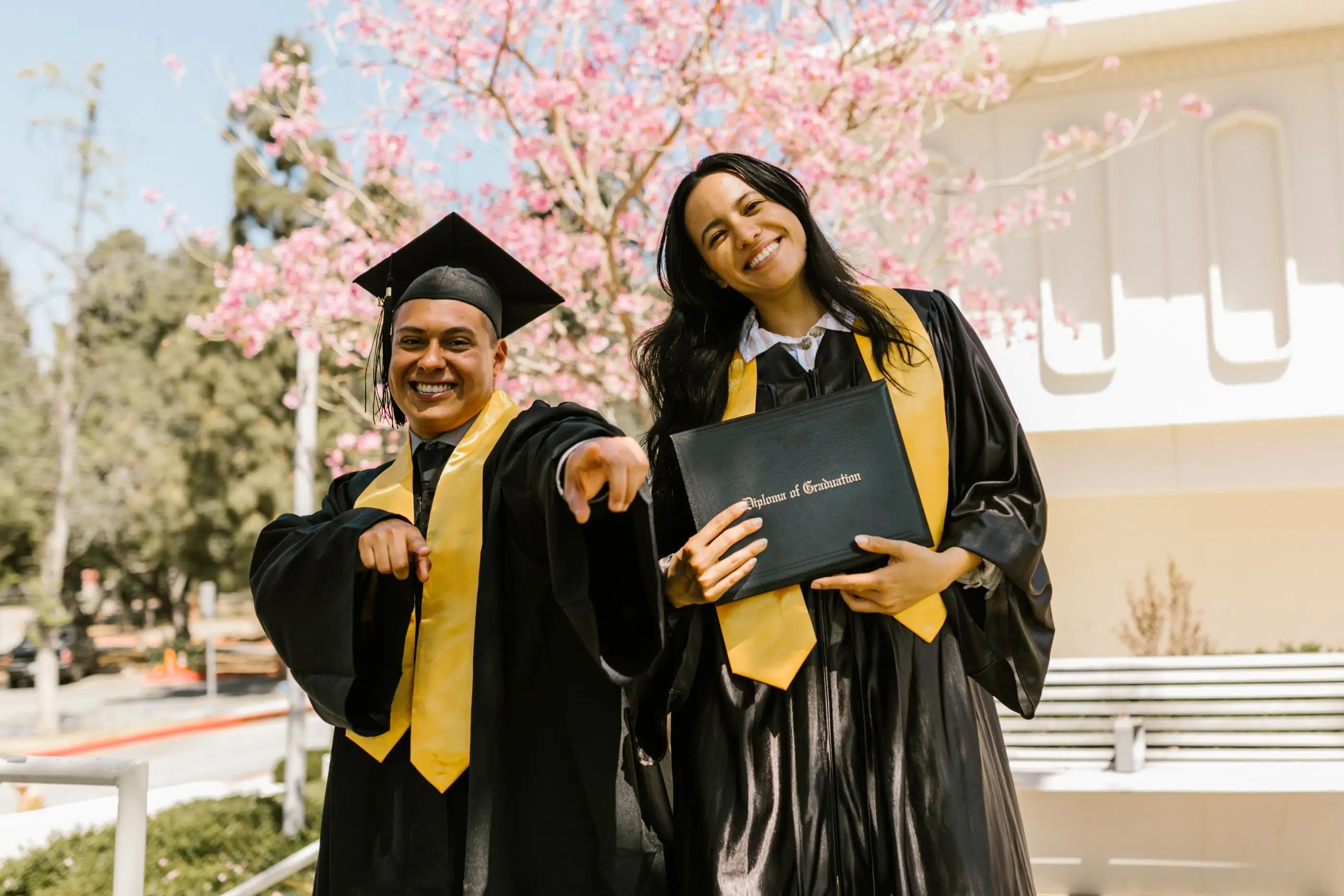 Two graduates in gowns smile and point toward the camera, holding their diploma folders.