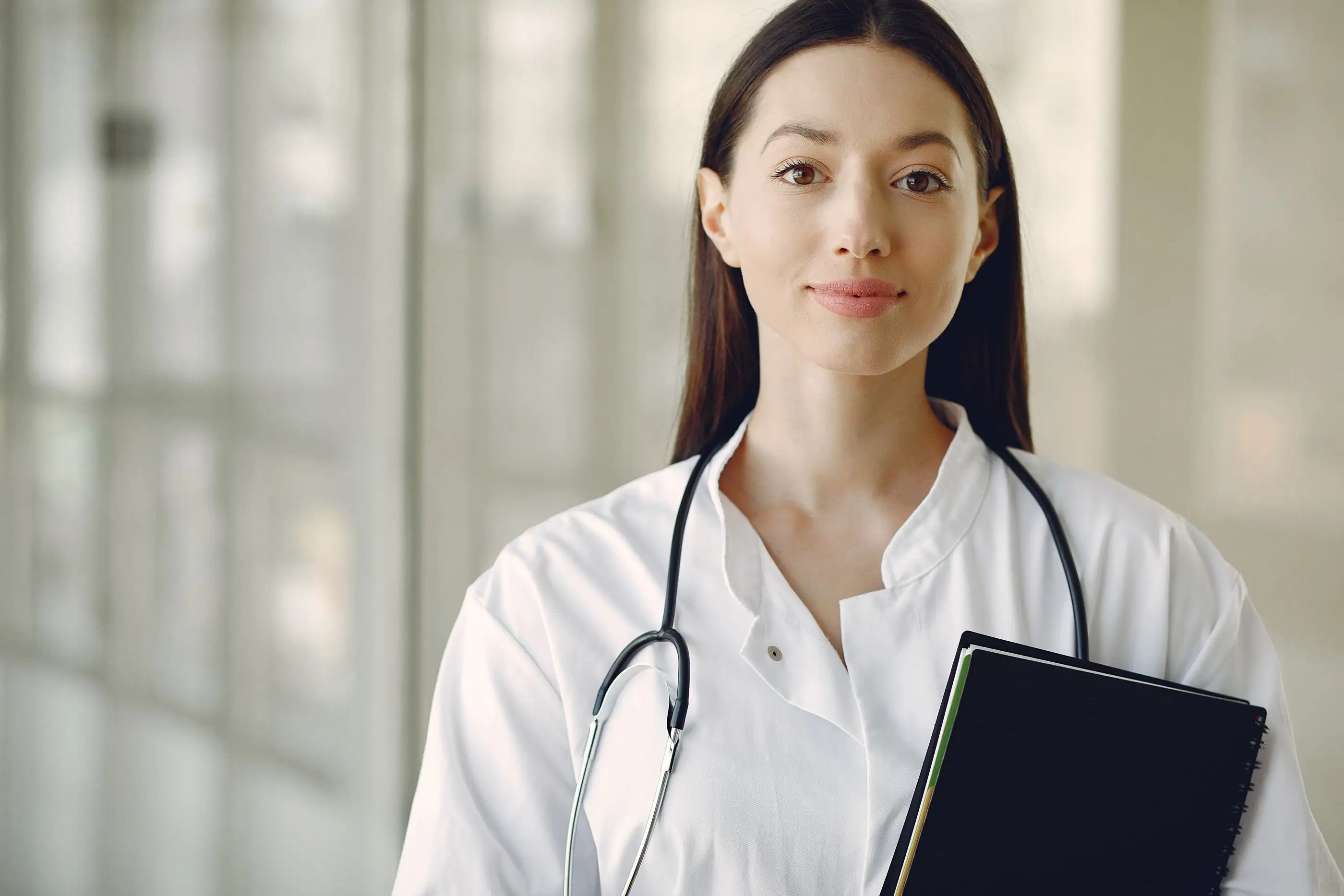 A smiling young clinician in a white coat holds a notebook in a bright room.
