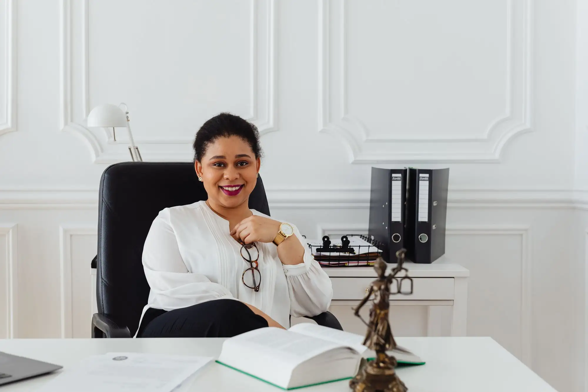 A smiling professional sits at her desk in a bright office with law books.