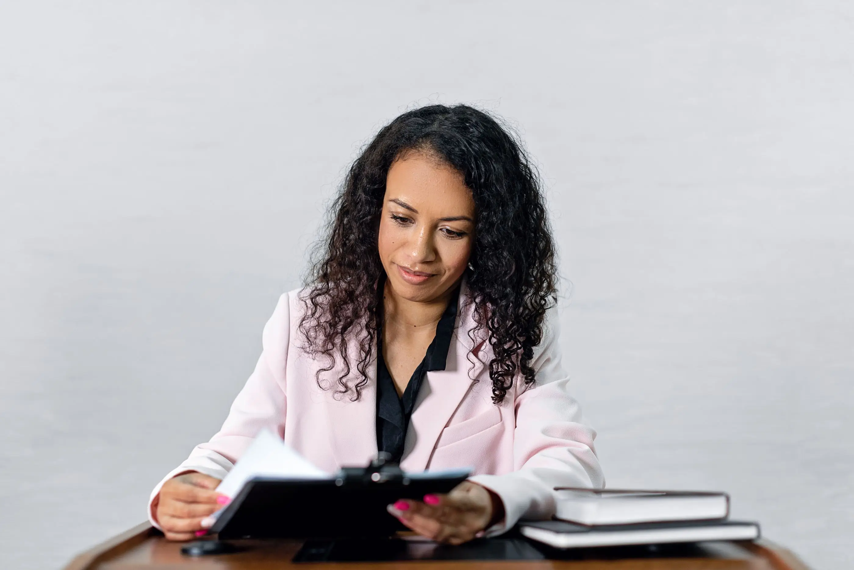 A woman in a light blazer reviews papers on a clipboard at her desk.