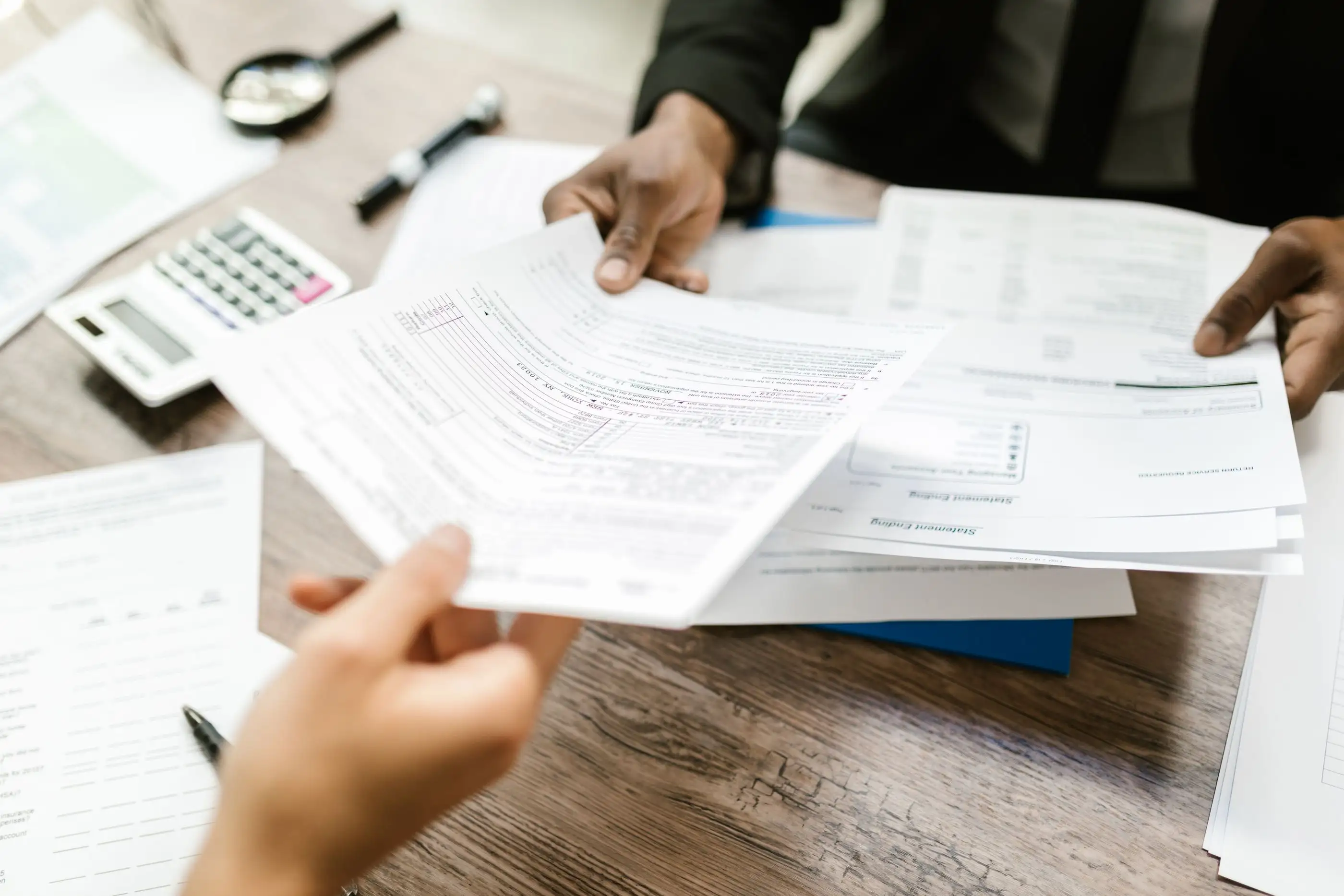 Two people pass application papers across a desk beside a calculator.