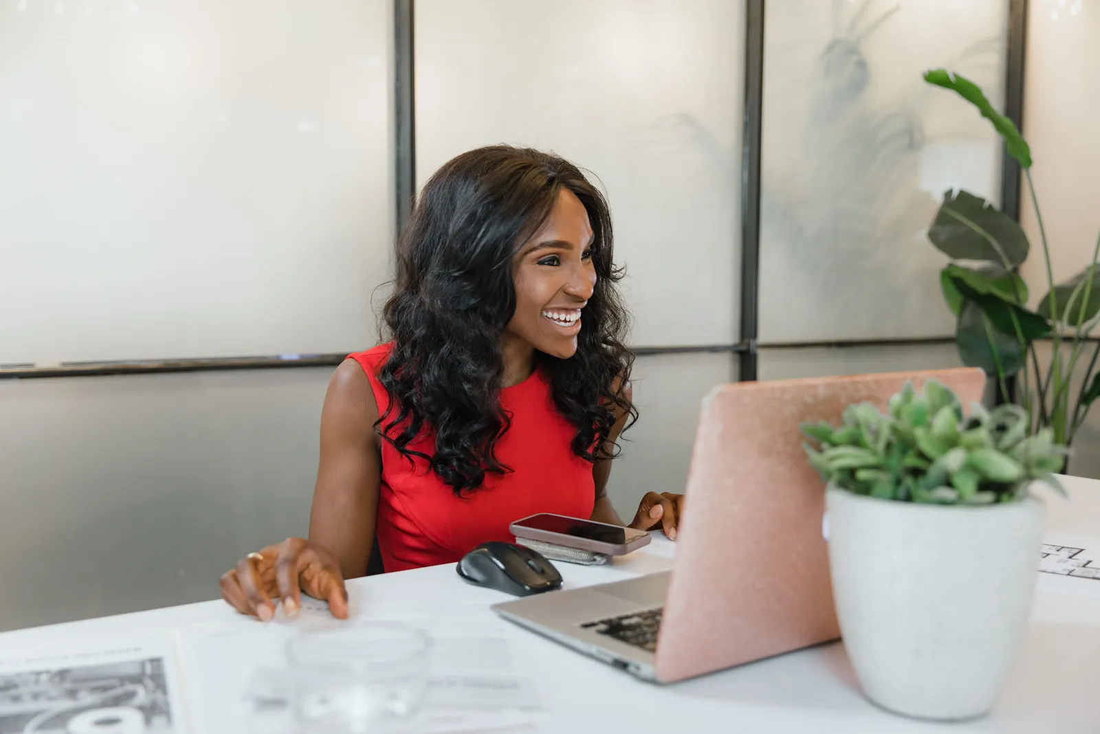 A smiling woman in a red top works at a laptop at a bright desk with plants.