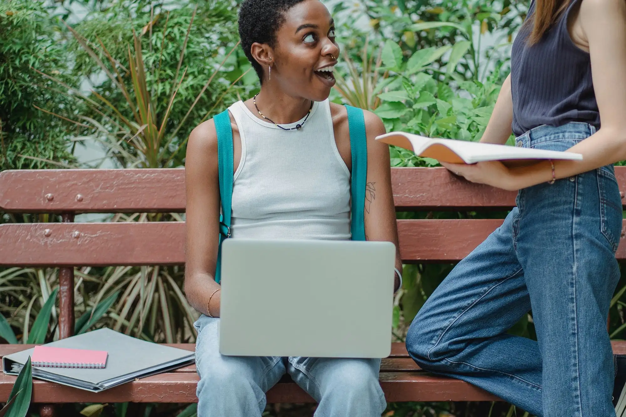 A student with a laptop on a park bench looks up, delighted, at a friend holding a notebook.
