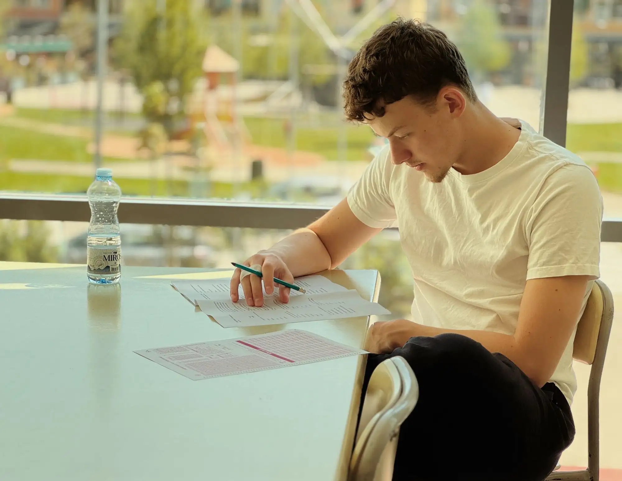 A student writes at a table beside a bright window.