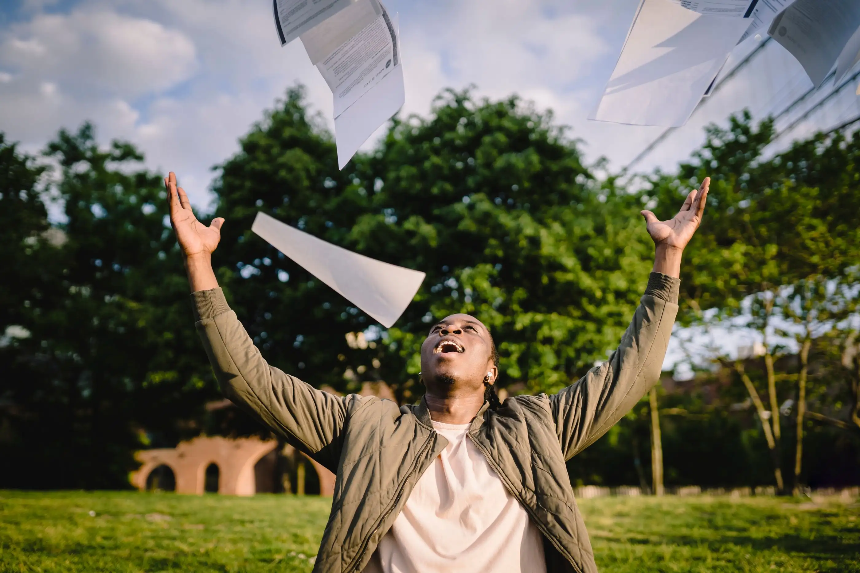 A student in a park throws his papers into the air in celebration.