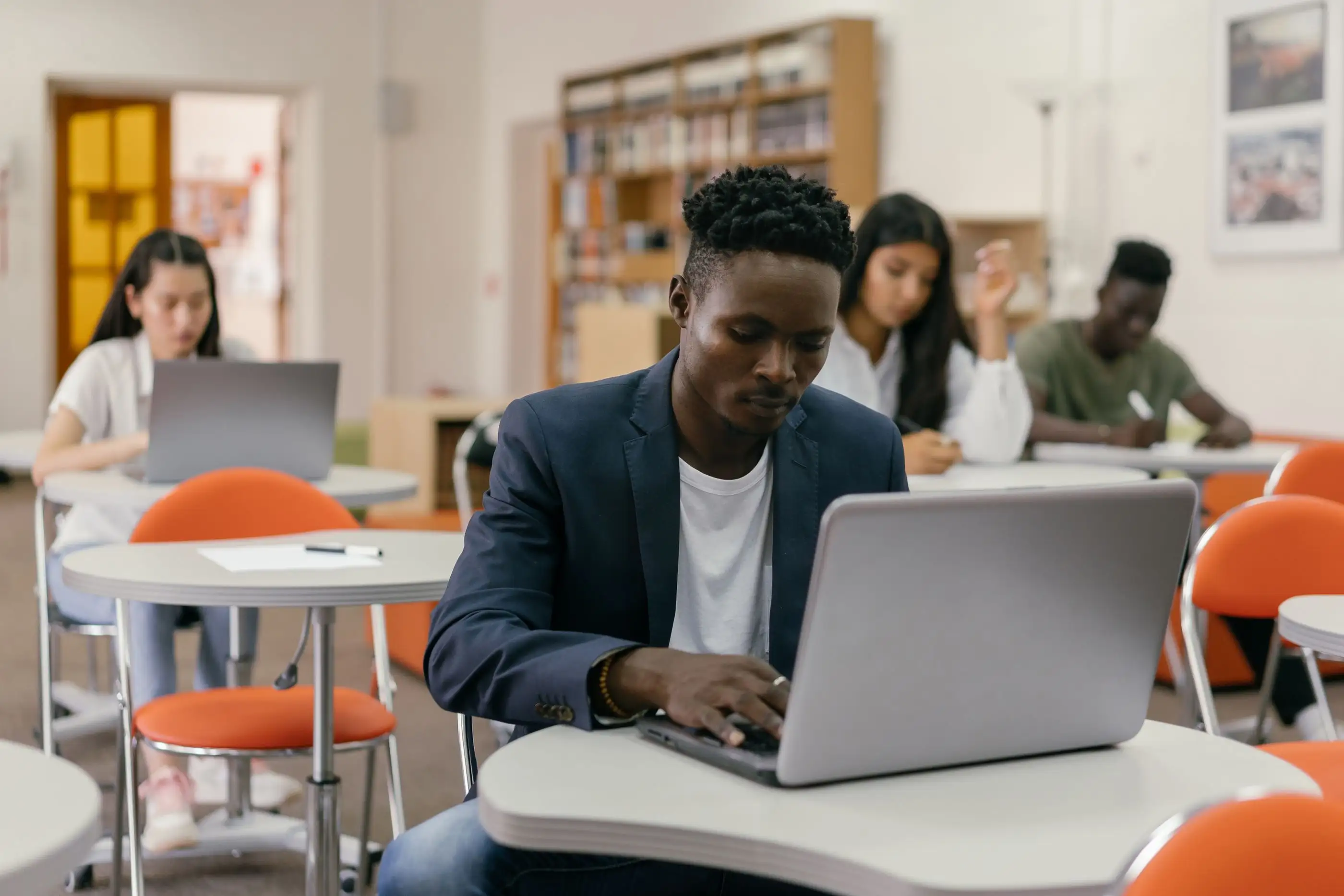 A student works on a laptop at a desk while others study in the room behind him.