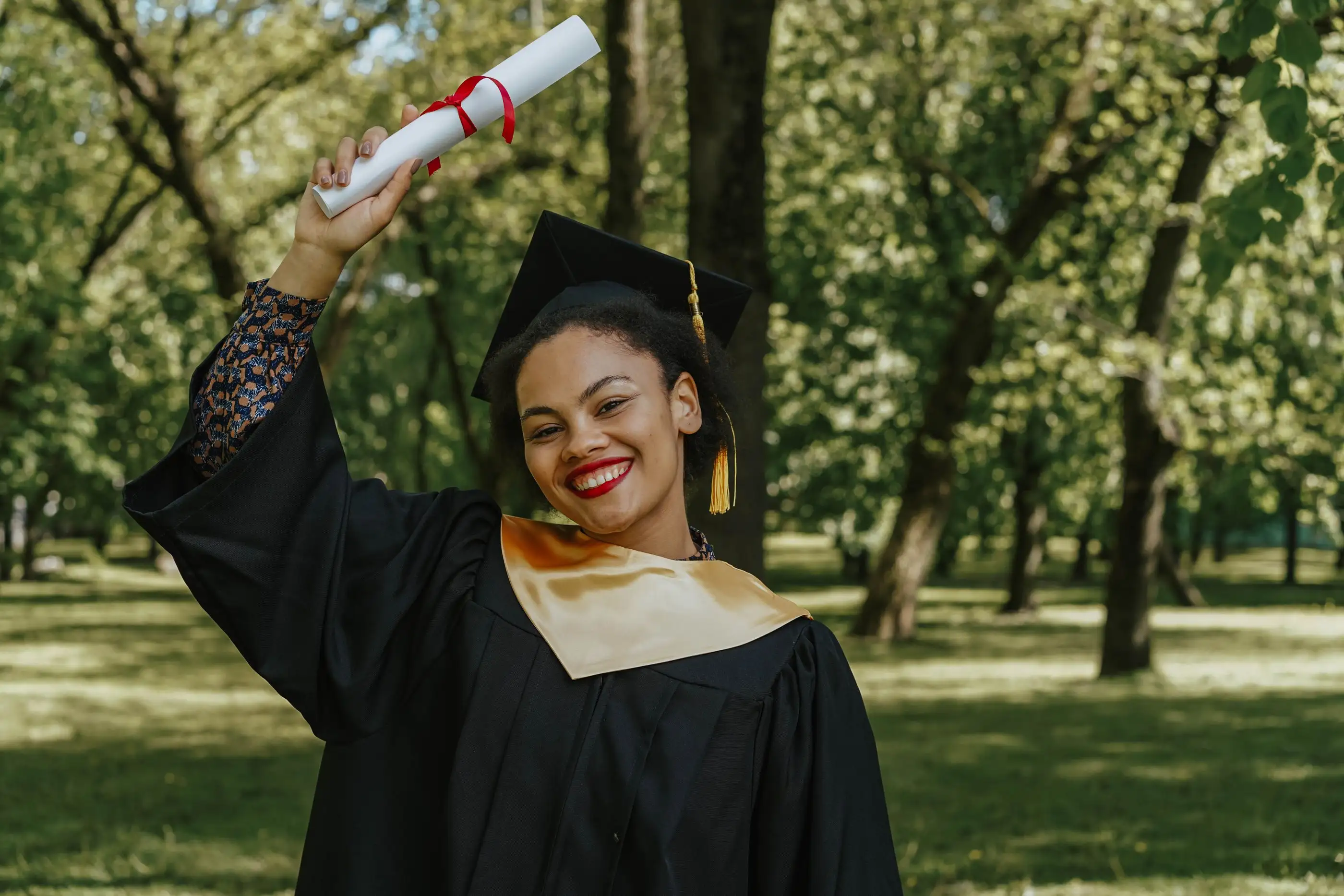 A graduate in a gown raises her diploma under the trees.