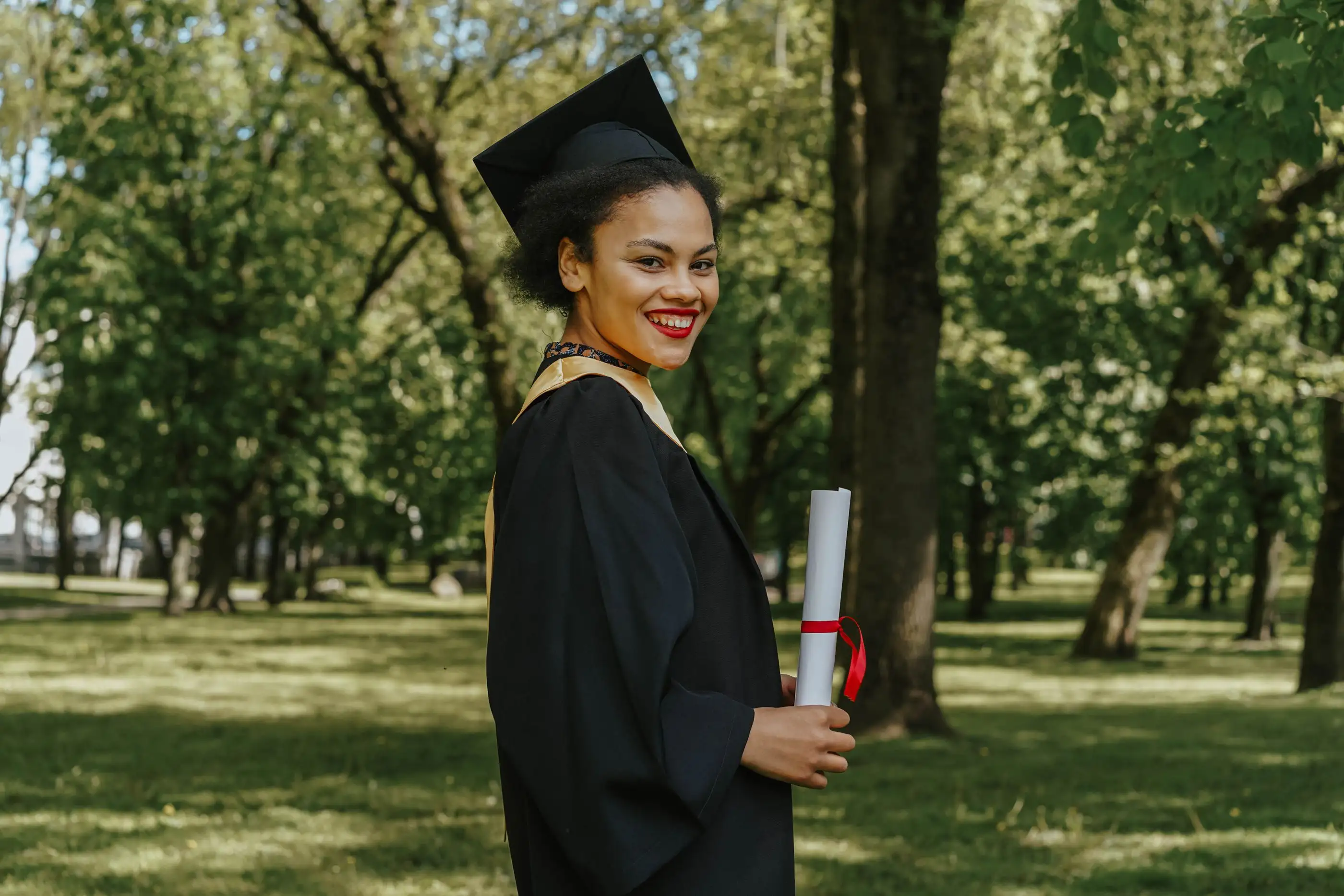 A smiling graduate in cap and gown holds her diploma in a park.