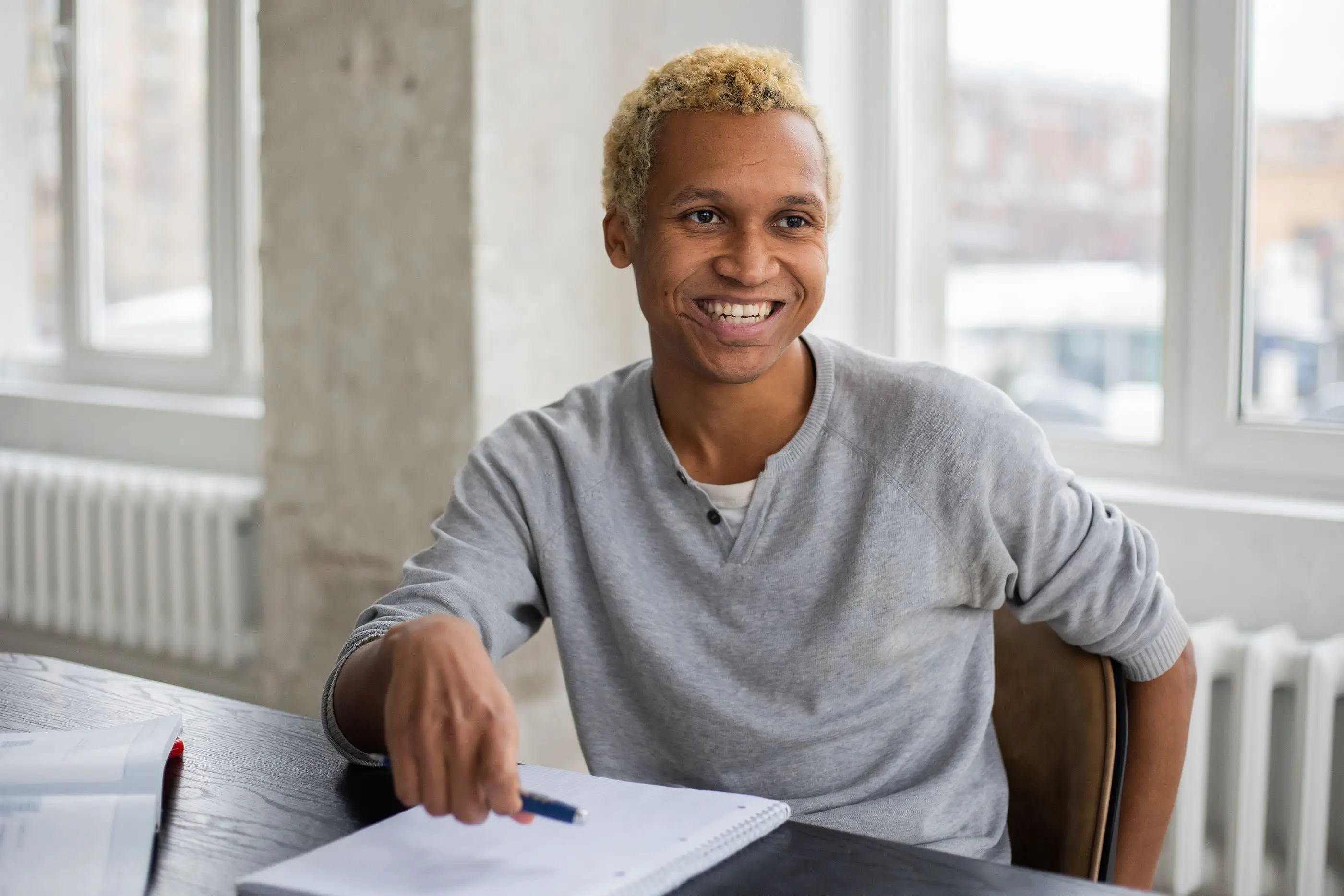 A smiling student sits at a desk with notes in a bright, sunlit room.