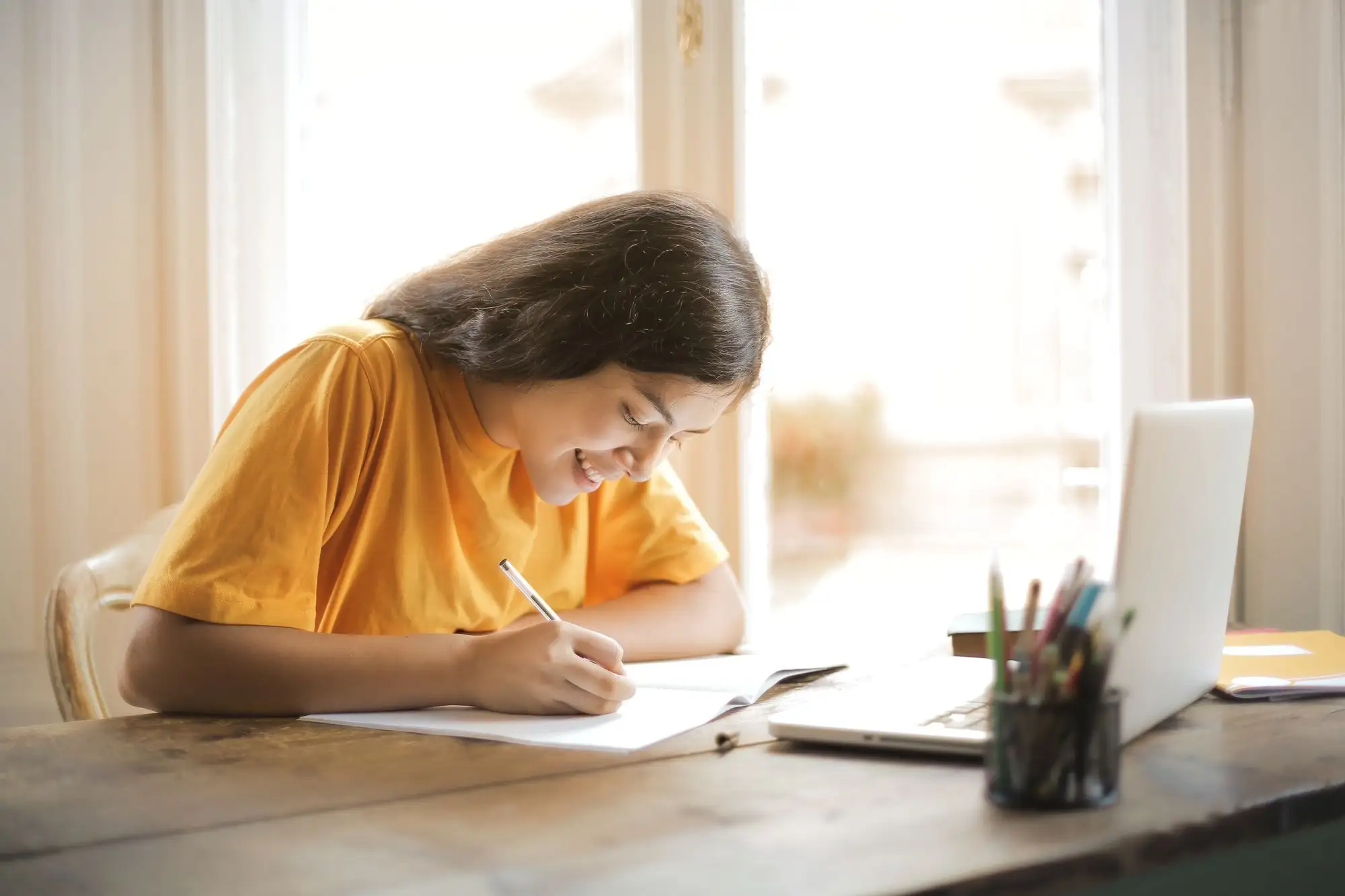 A student in a yellow shirt writes notes beside a laptop by a window.
