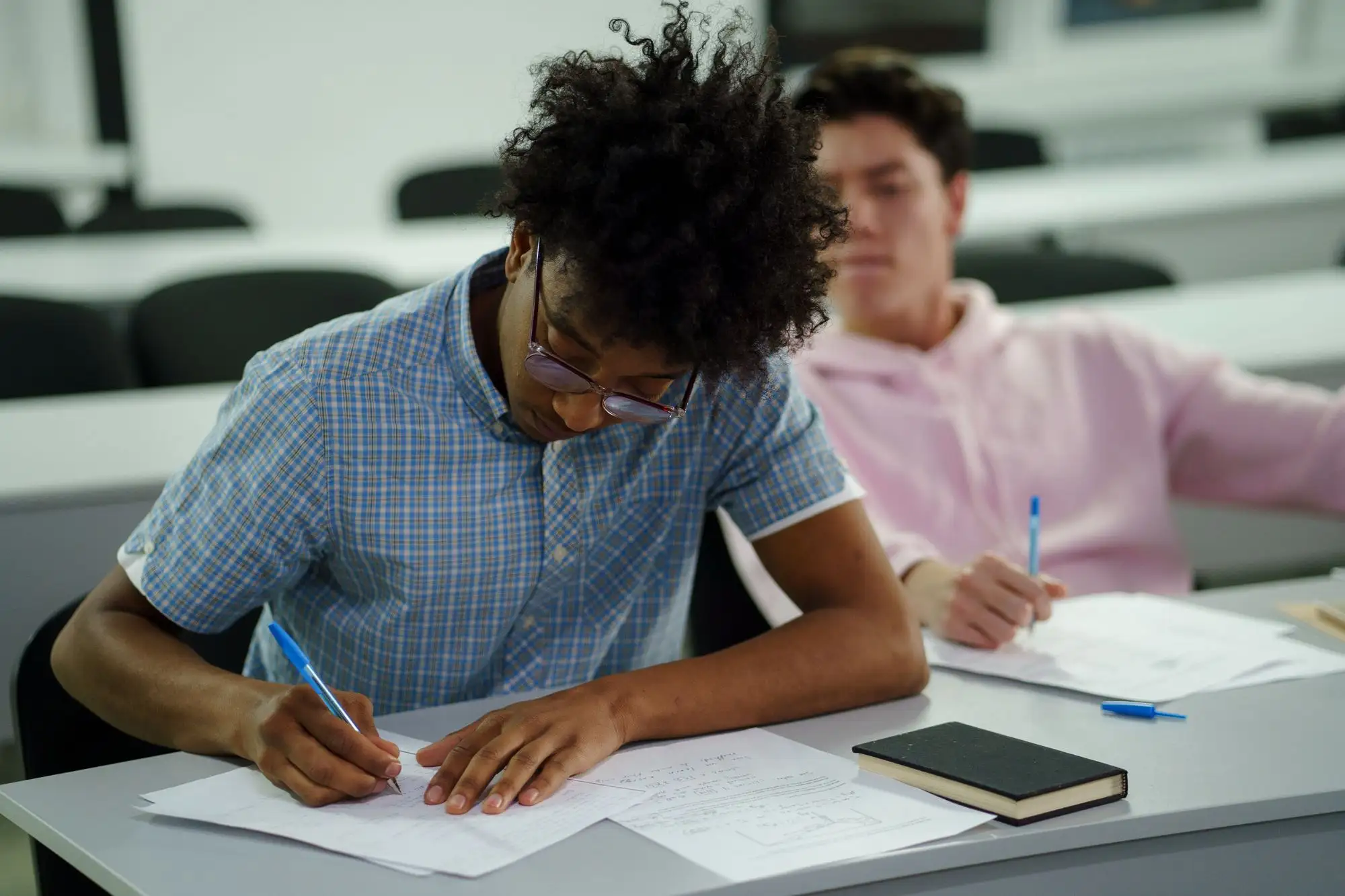 A student writes answers at a desk in a bright classroom.