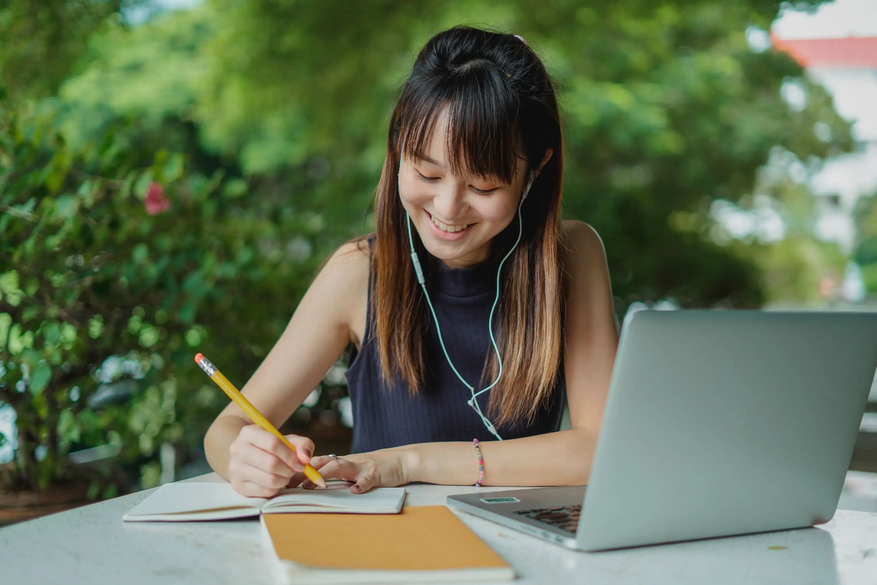A student smiles while writing notes beside her laptop outdoors.