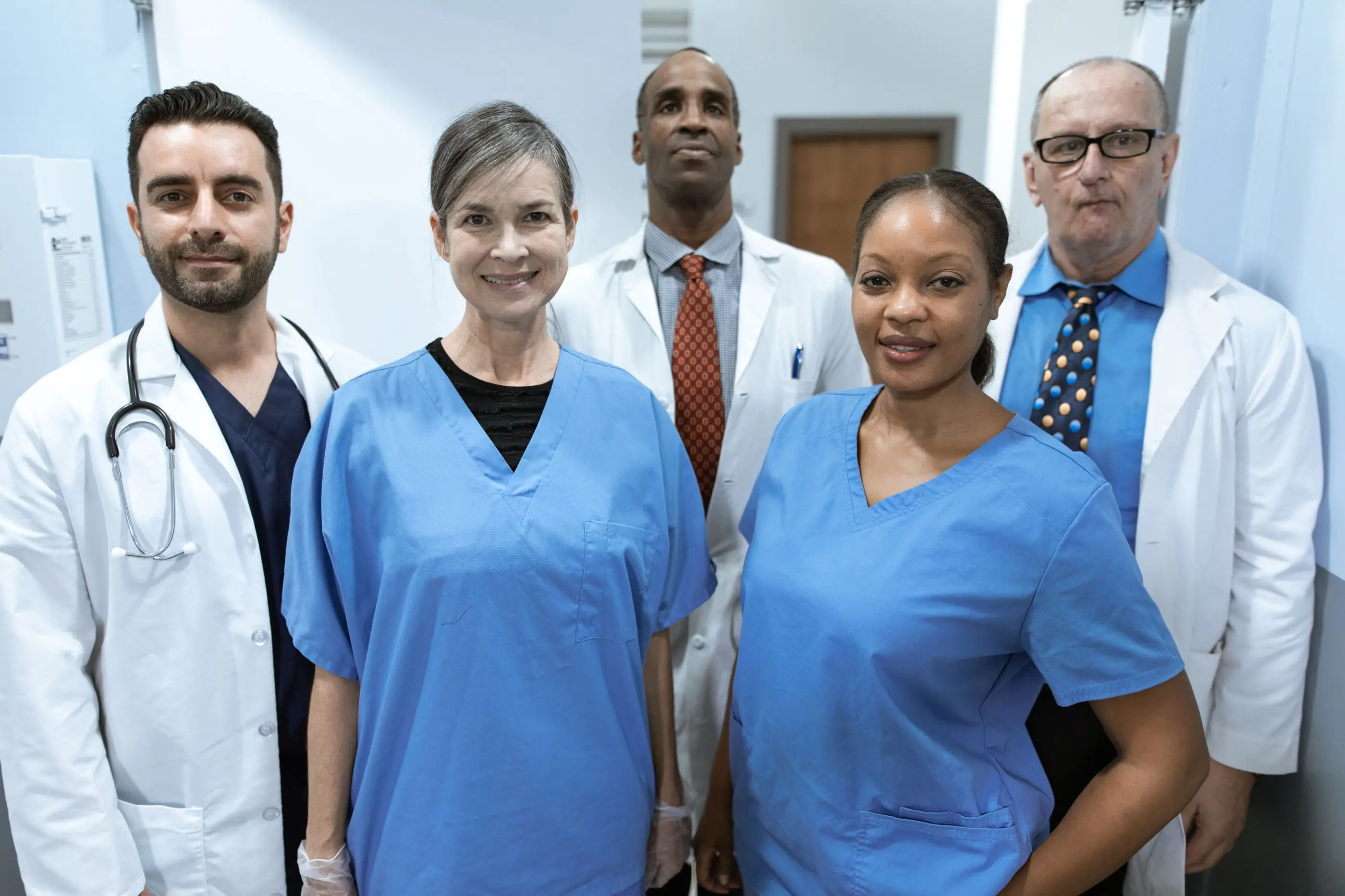 A smiling team of clinicians in white coats and scrubs stands together in a hospital corridor.