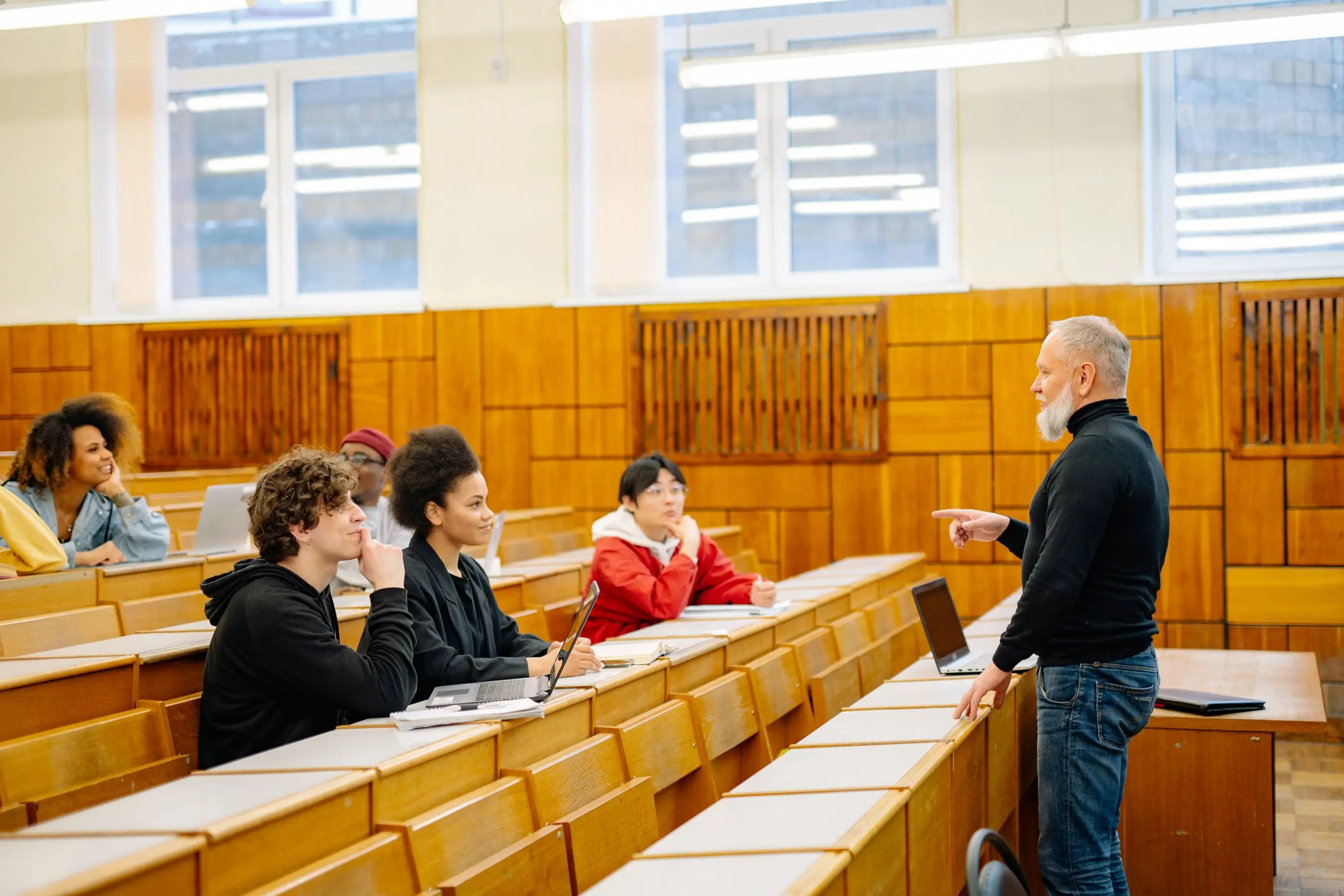 An instructor talks with students in a sunlit lecture hall.