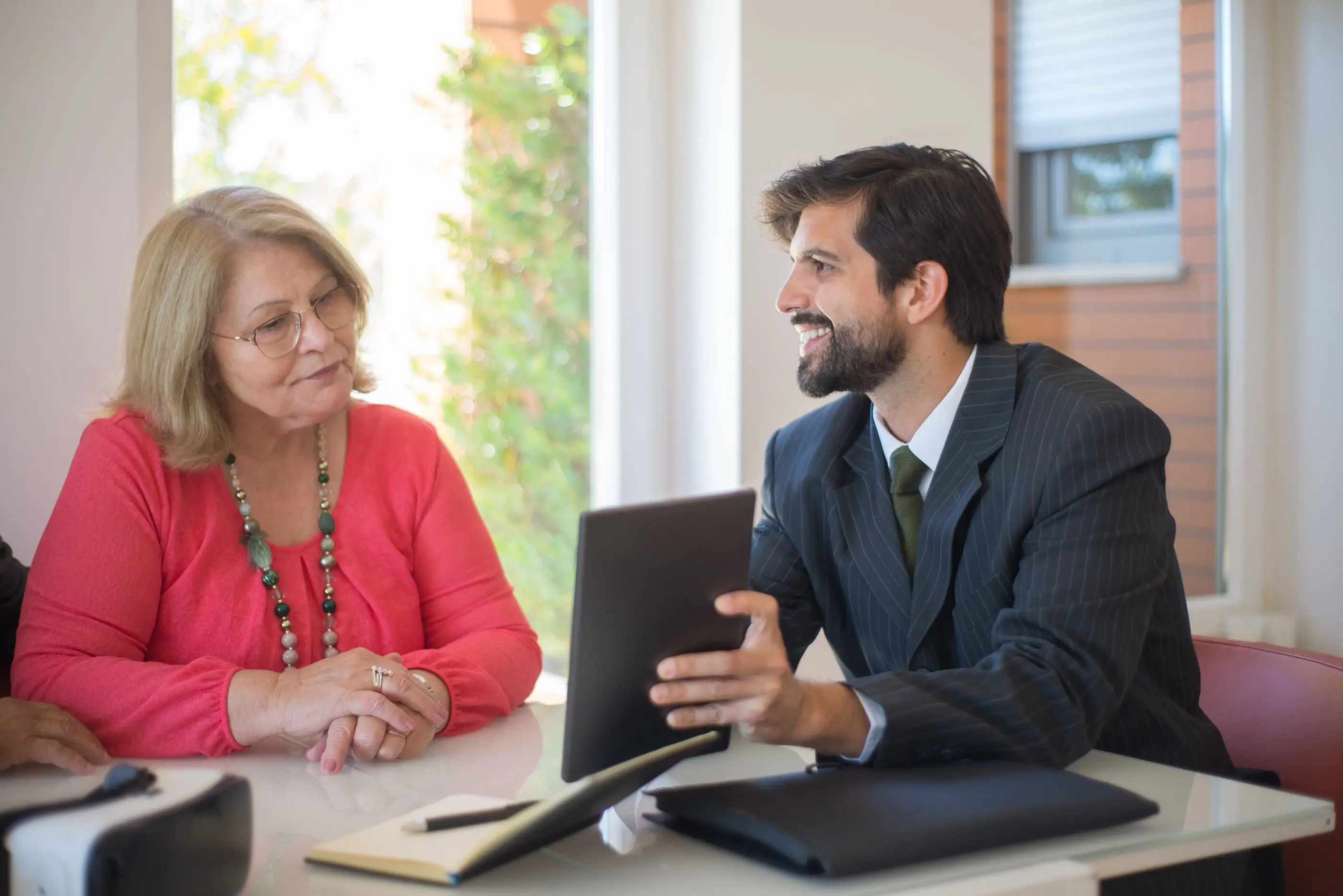 An adviser talks an older woman through information on a tablet at a bright table.